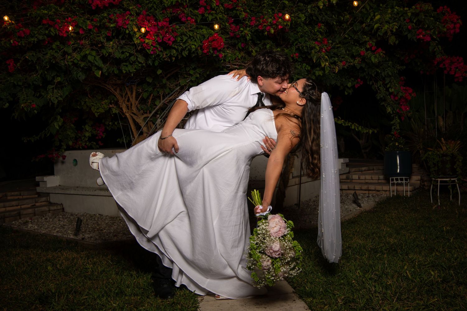 A romantic dip pose between newlyweds in front of a garden with red flowers and twinkling lights. Wedding photography.