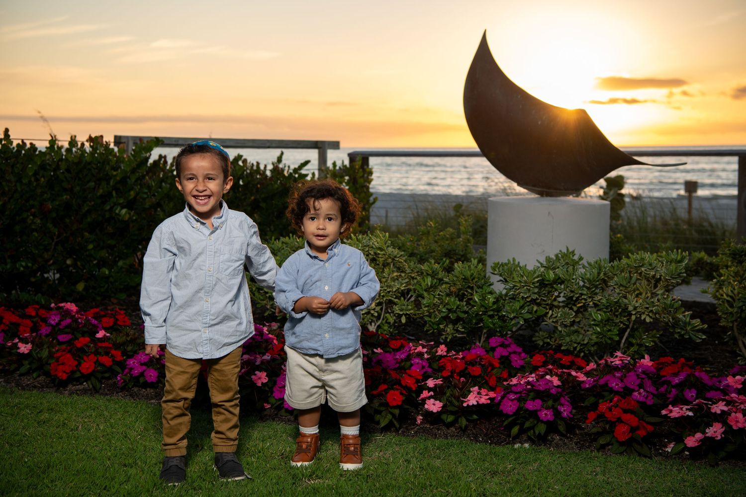 Two young children stand in front of a sunset garden with pink flowers and a curved sculpture. Family photoshoot.