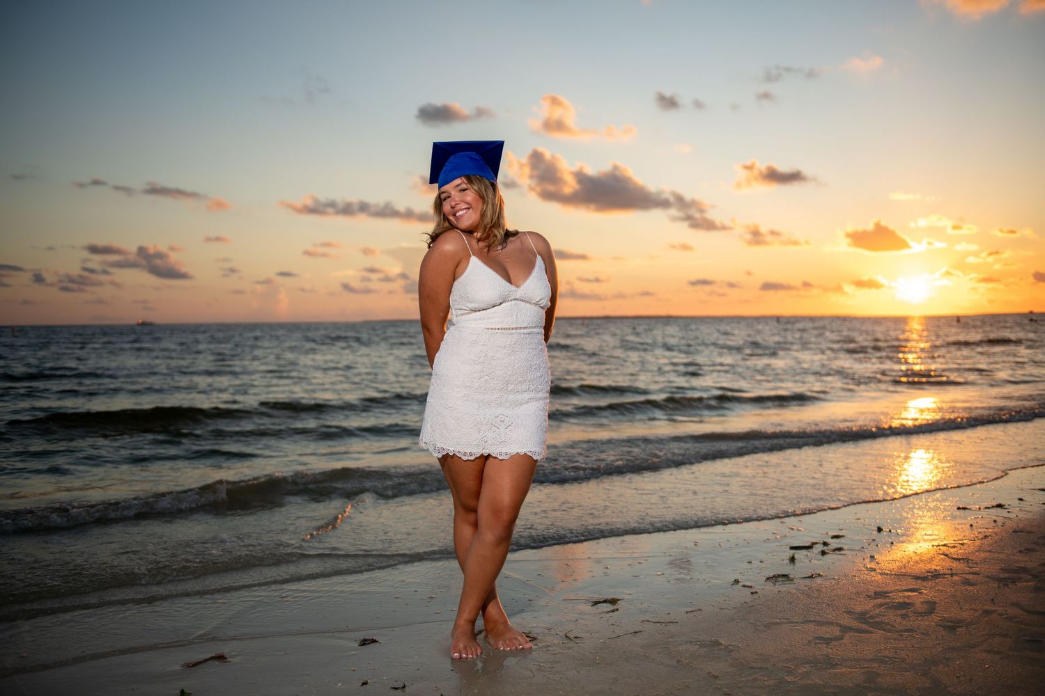 Graduate in white dress and blue cap poses on beach during golden sunset.