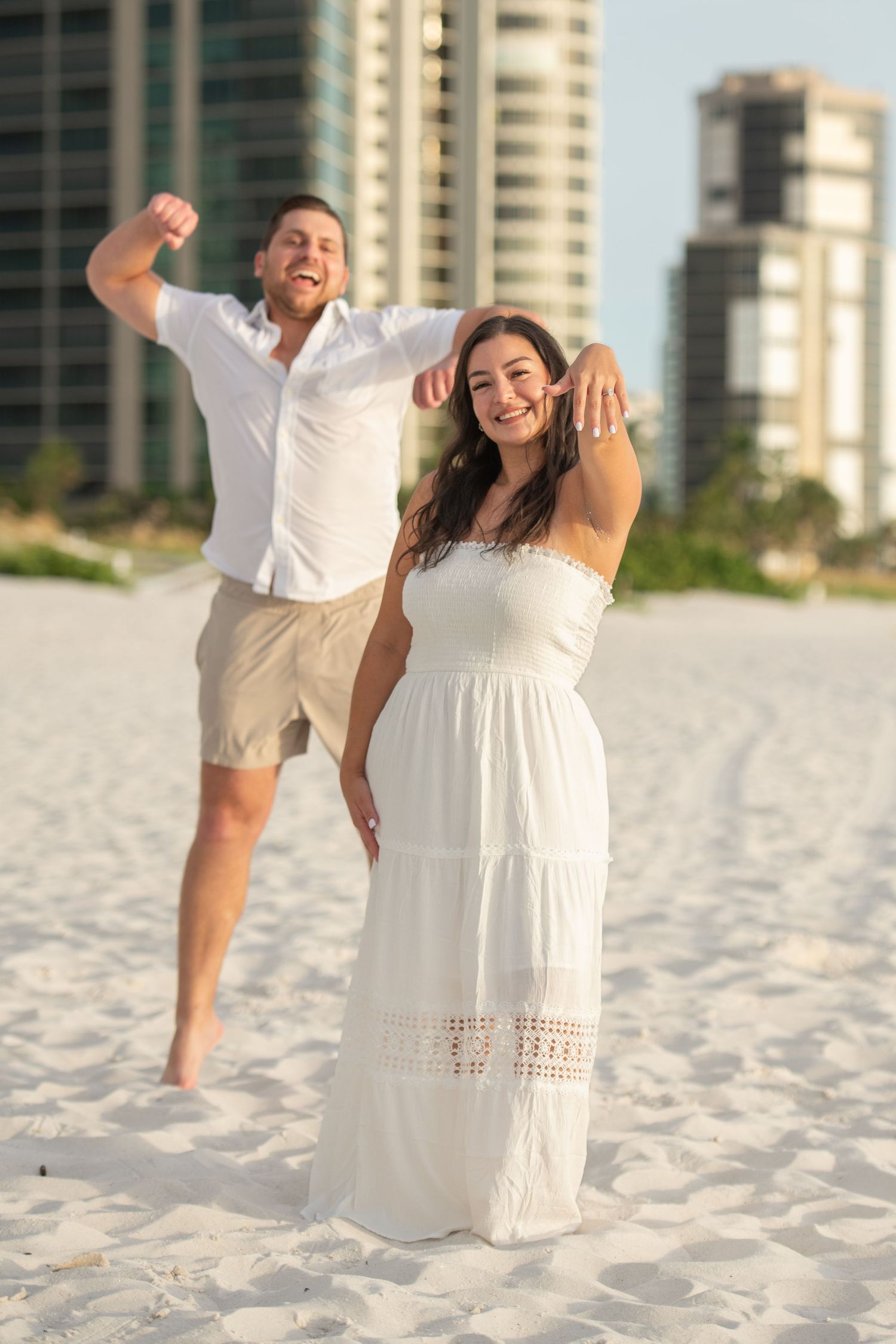A couple shares joyful moments together on a white sandy beach with high-rise buildings in the background.