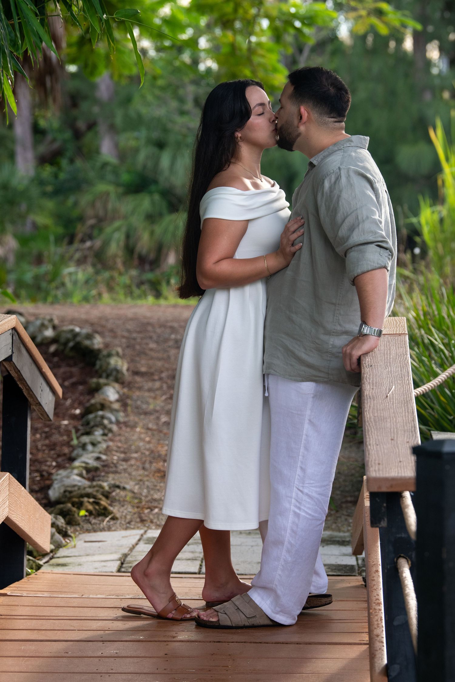A romantic couple in white and gray outfits share tender moments on a wooden boardwalk surrounded by greenery.