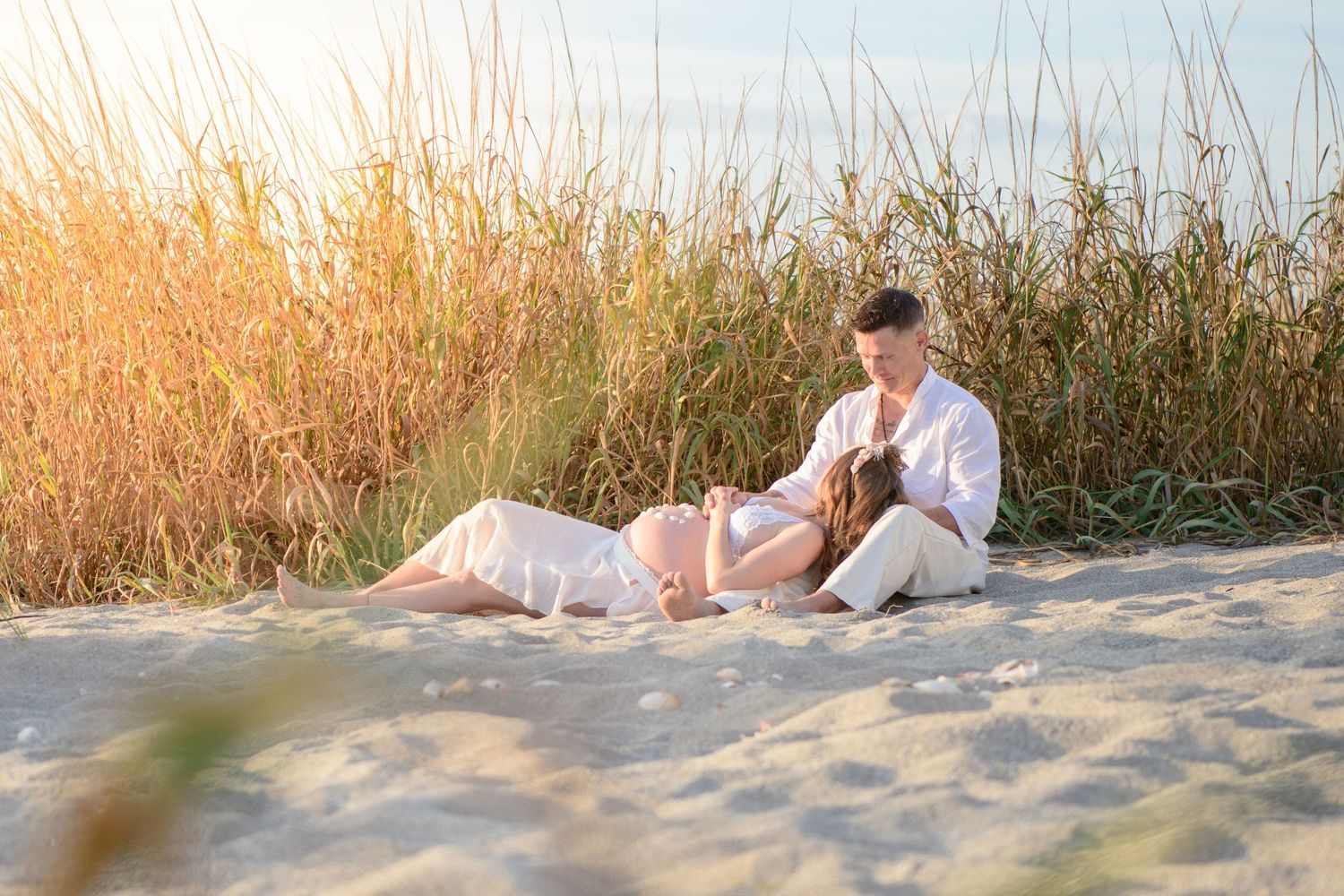 Couple lying in beach grass during golden hour photoshoot. Maternity photoshoot,