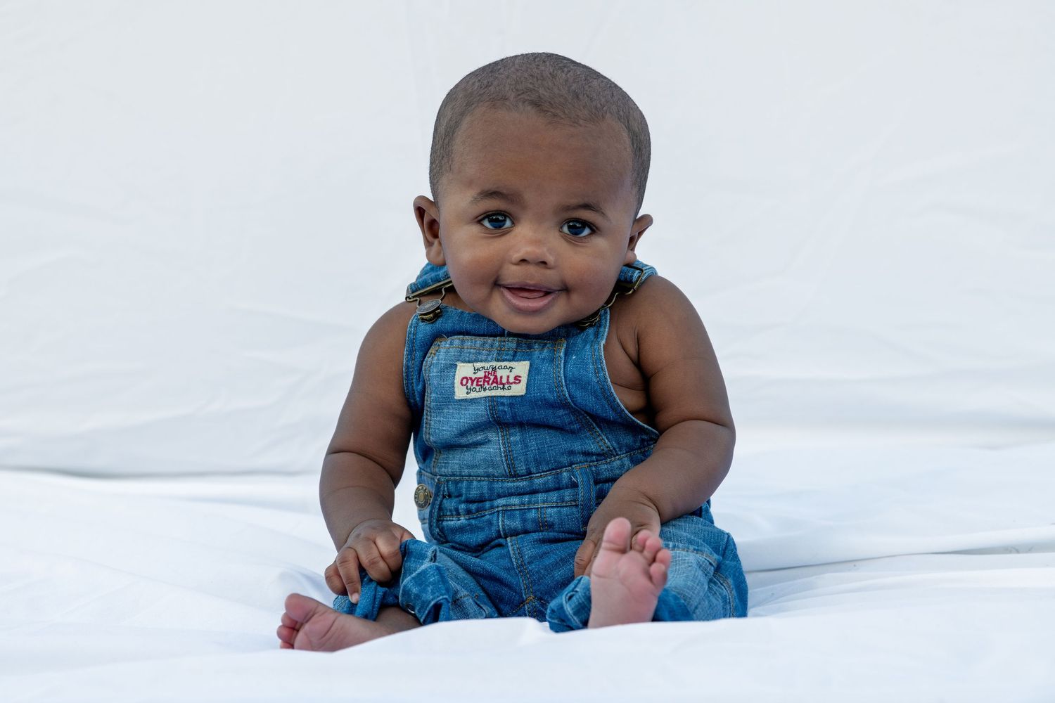 Baby wearing blue denim overalls sits on white surface.