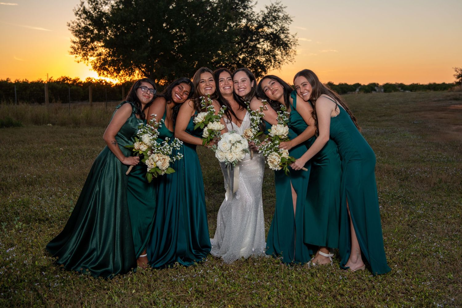 Bridal party in emerald dresses poses casually on grass during sunset photo session. Wedding photography.