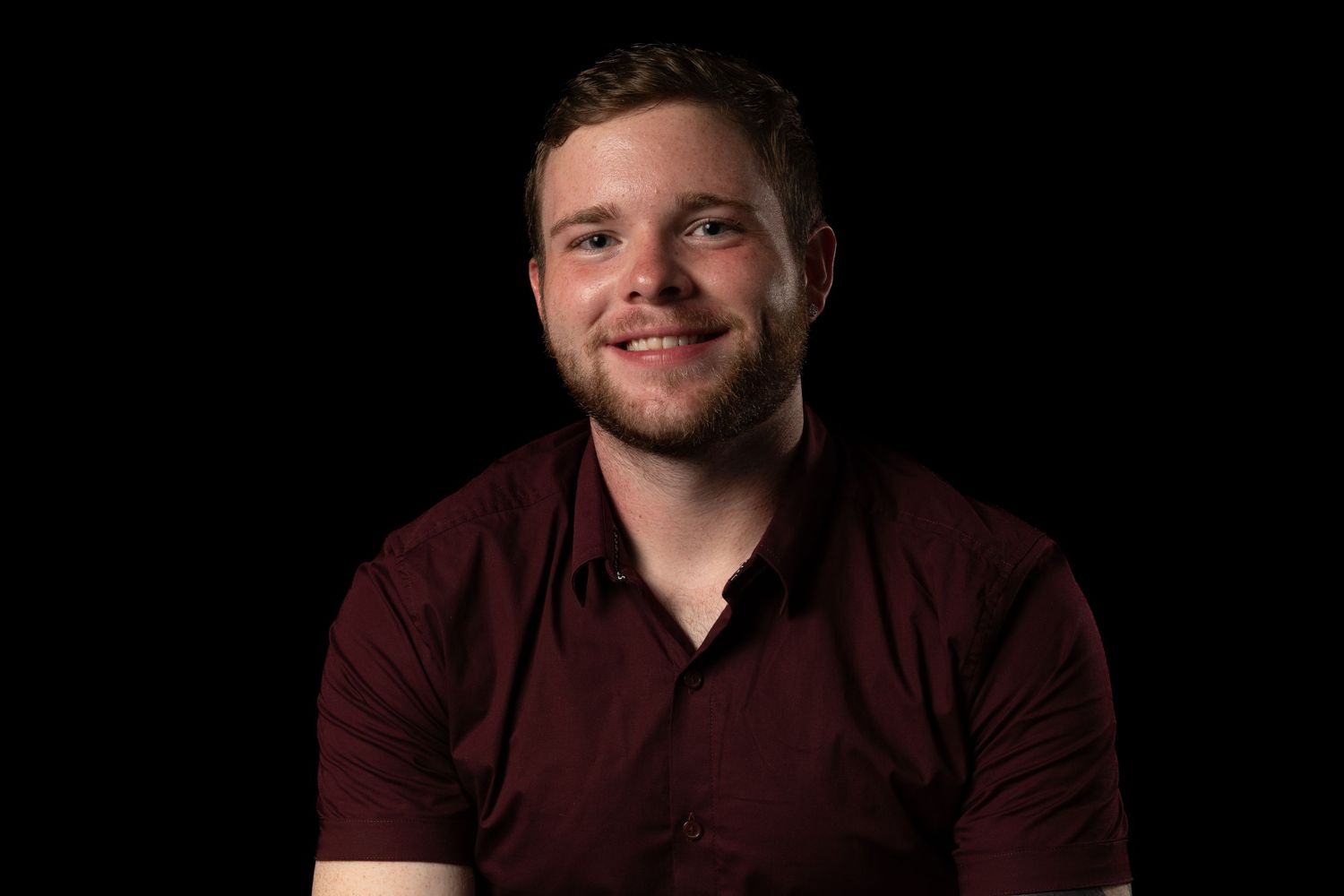 Professional headshot of person in maroon shirt against dark background.