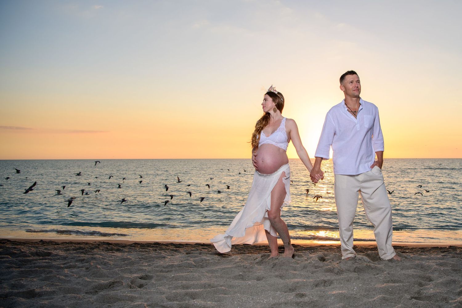 Beach photography session with white attire against setting sun and ocean waves. Maternity photoshoot,