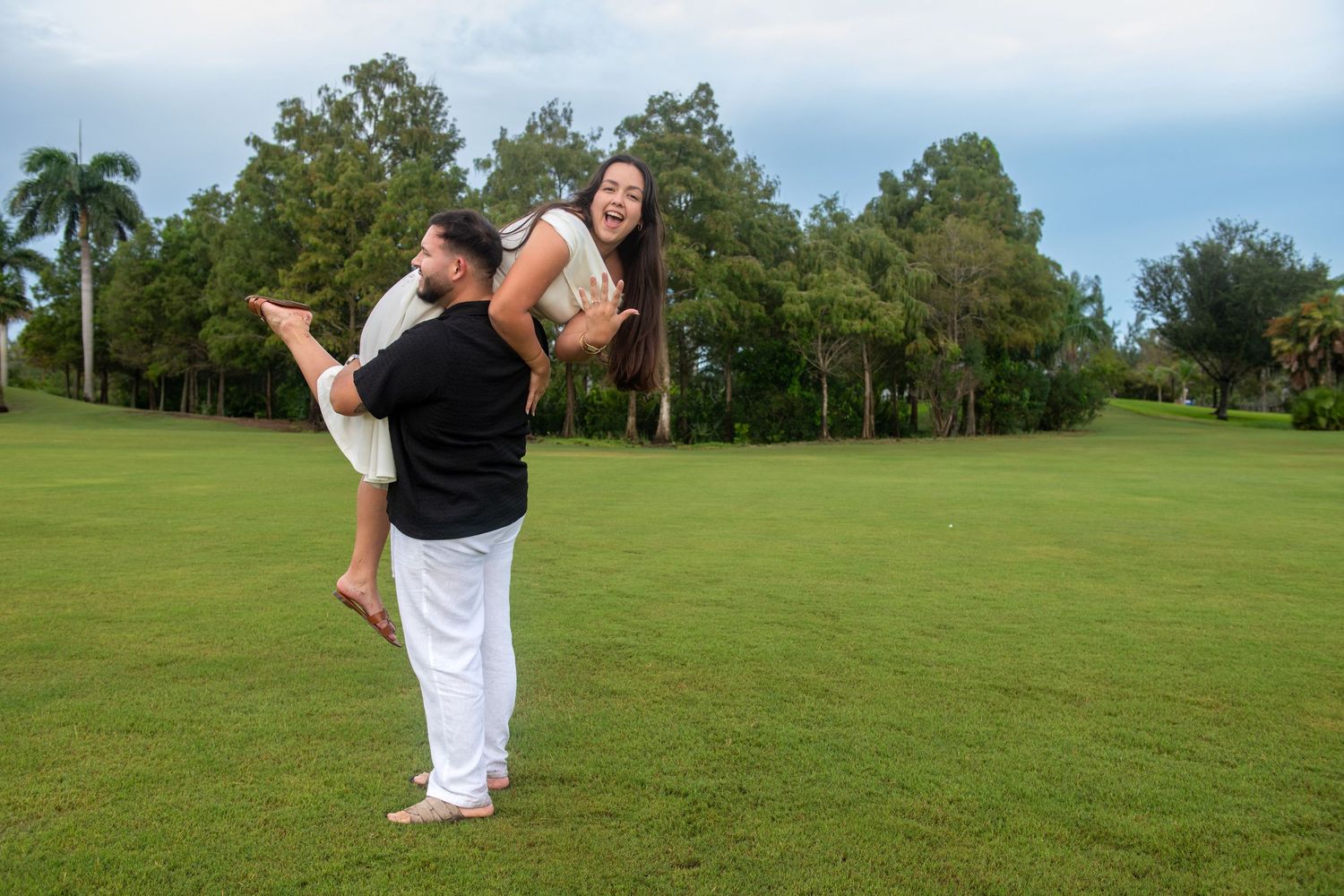 A couple poses playfully on a grassy field with palm trees in the background.