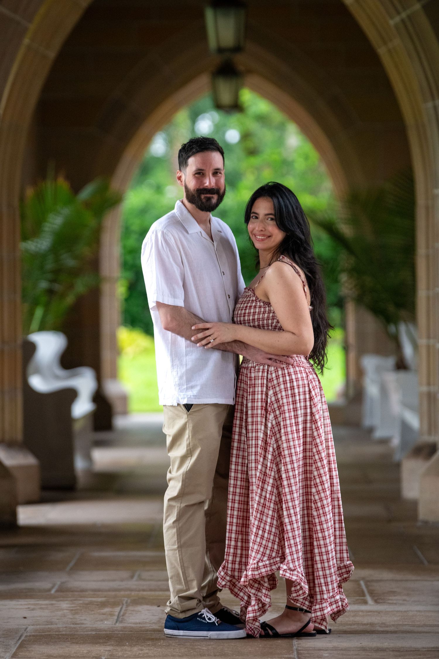 A couple embraces in a covered stone walkway with arched columns and greenery.