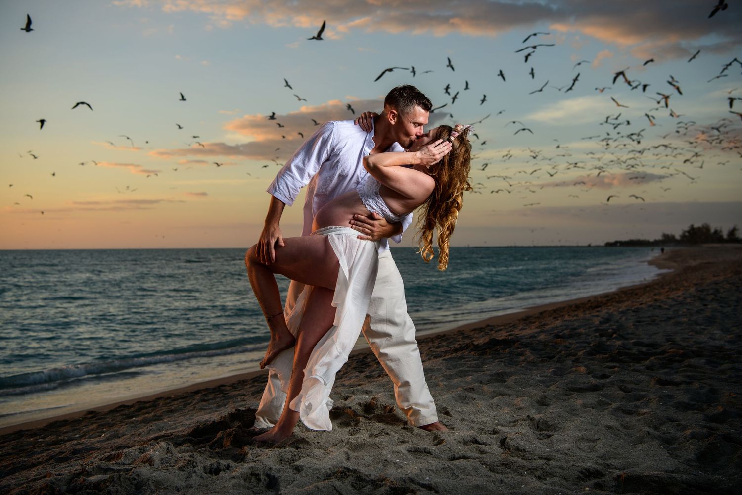 Couple performing romantic dance moves on beach with seabirds flying overhead at sunset. Maternity photoshoot,