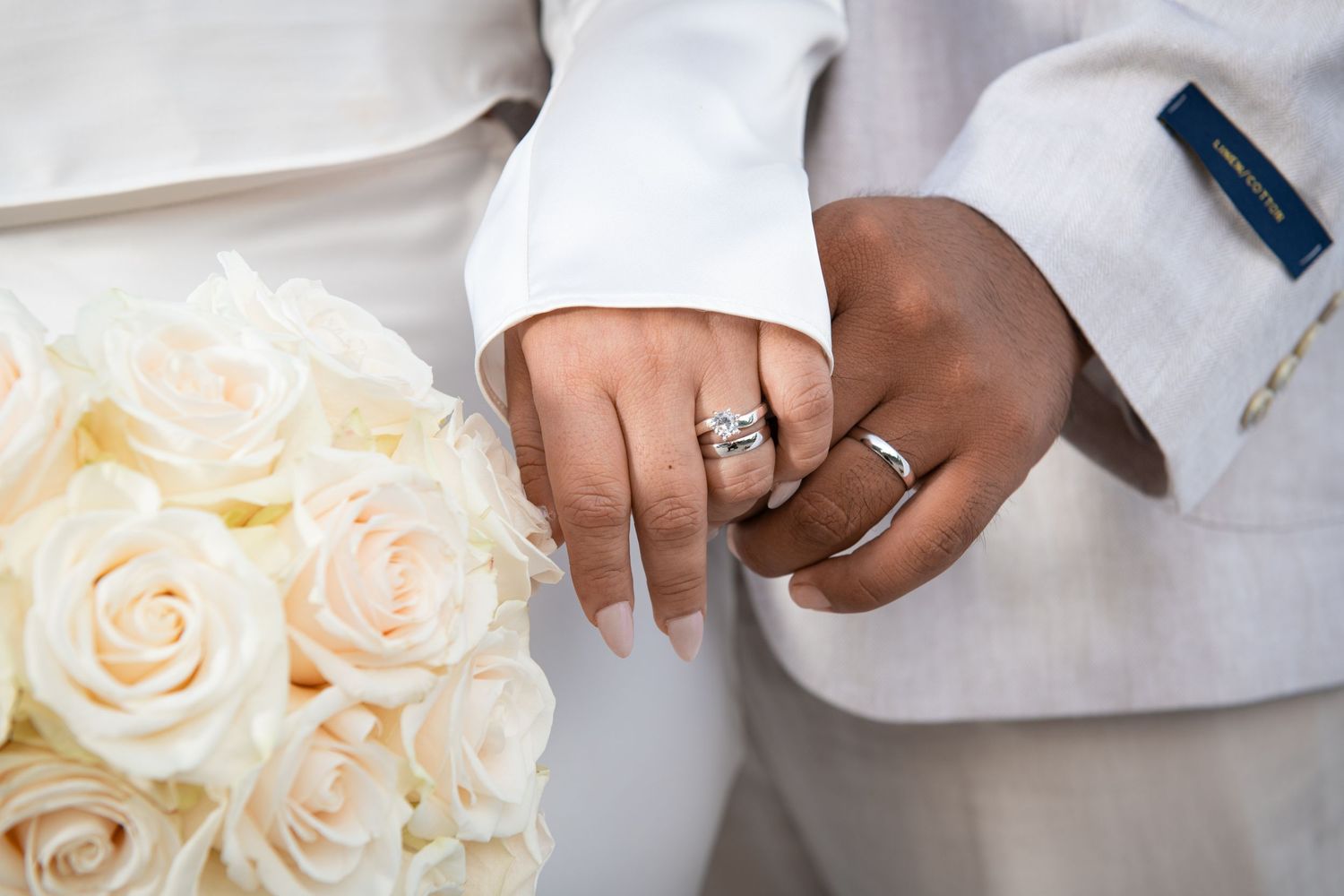 Close-up of hands wearing wedding rings with white roses in background. Wedding photography.