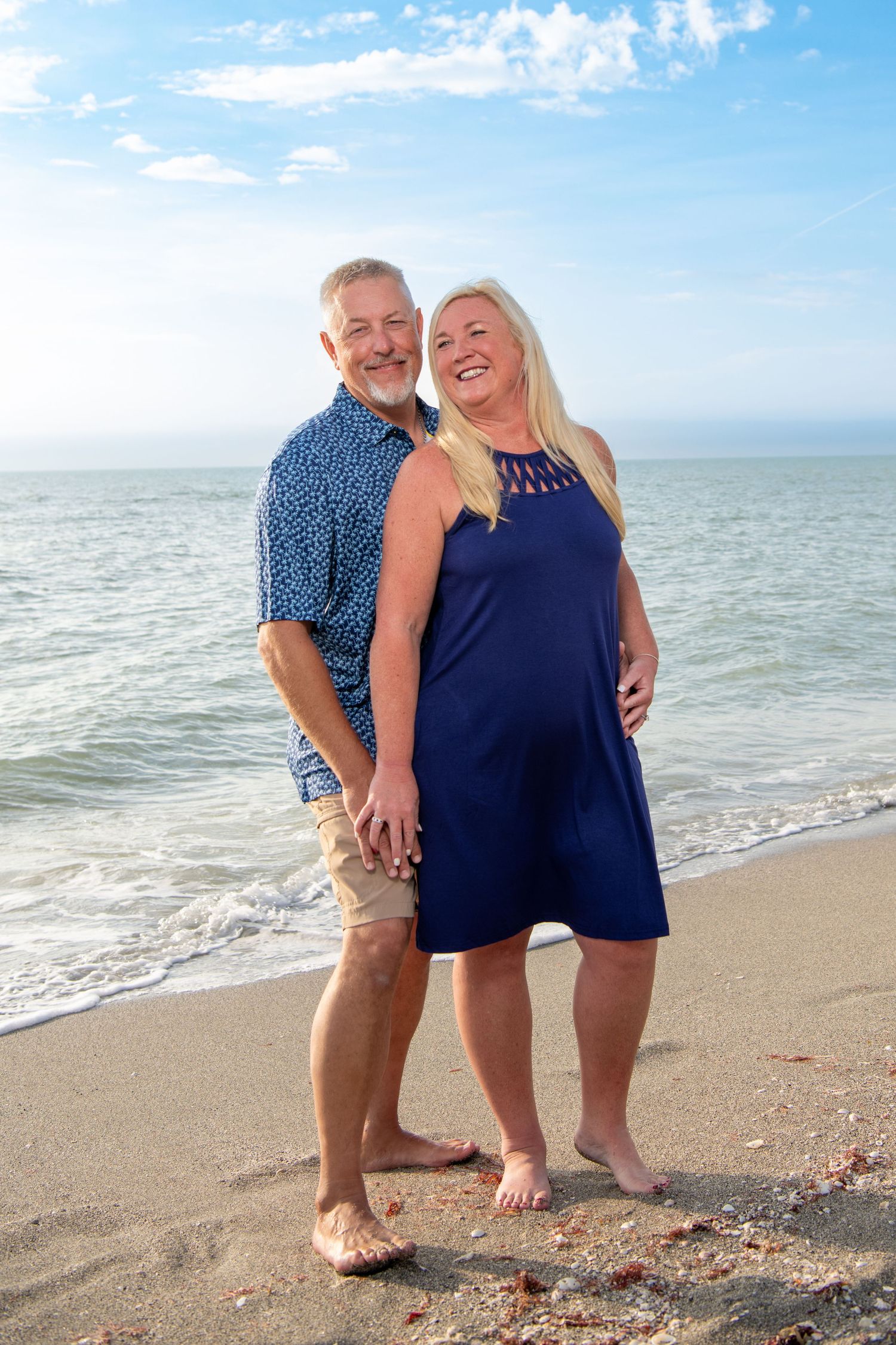 Couple in coordinating navy blue outfits walking barefoot along a sunny beach shoreline. Family photoshoot.