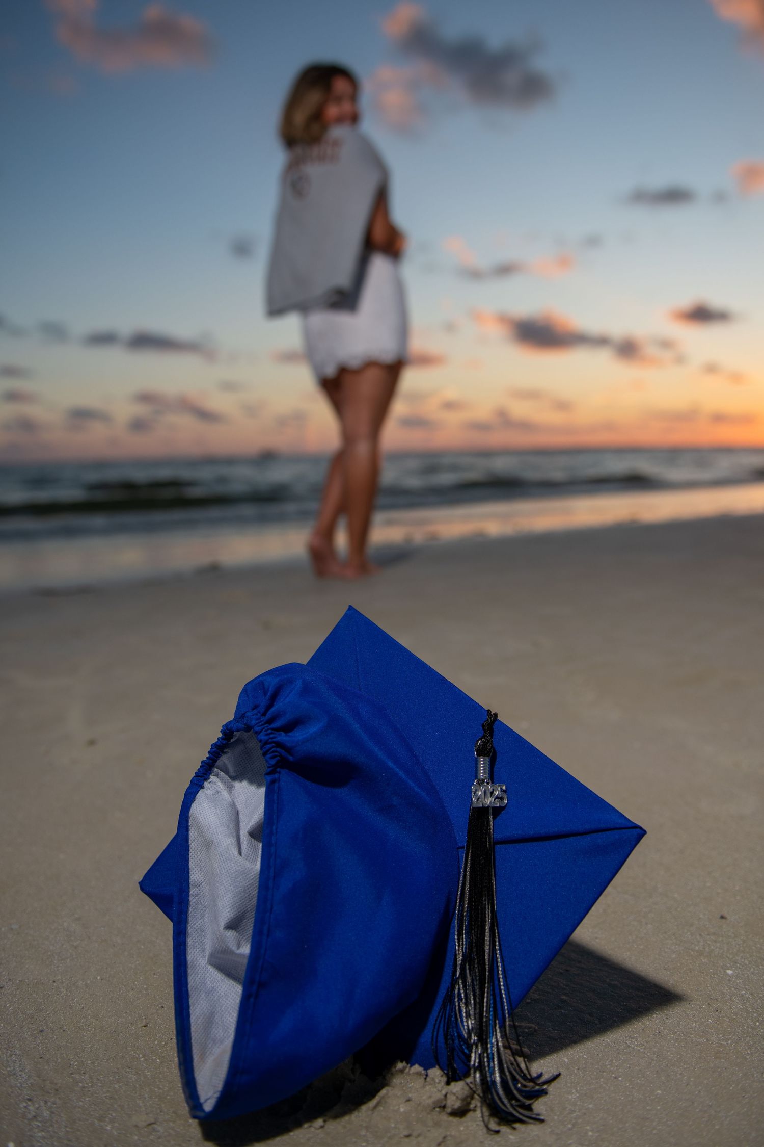 Blue graduation cap with tassel lying on beach sand with blurred figure in background.
