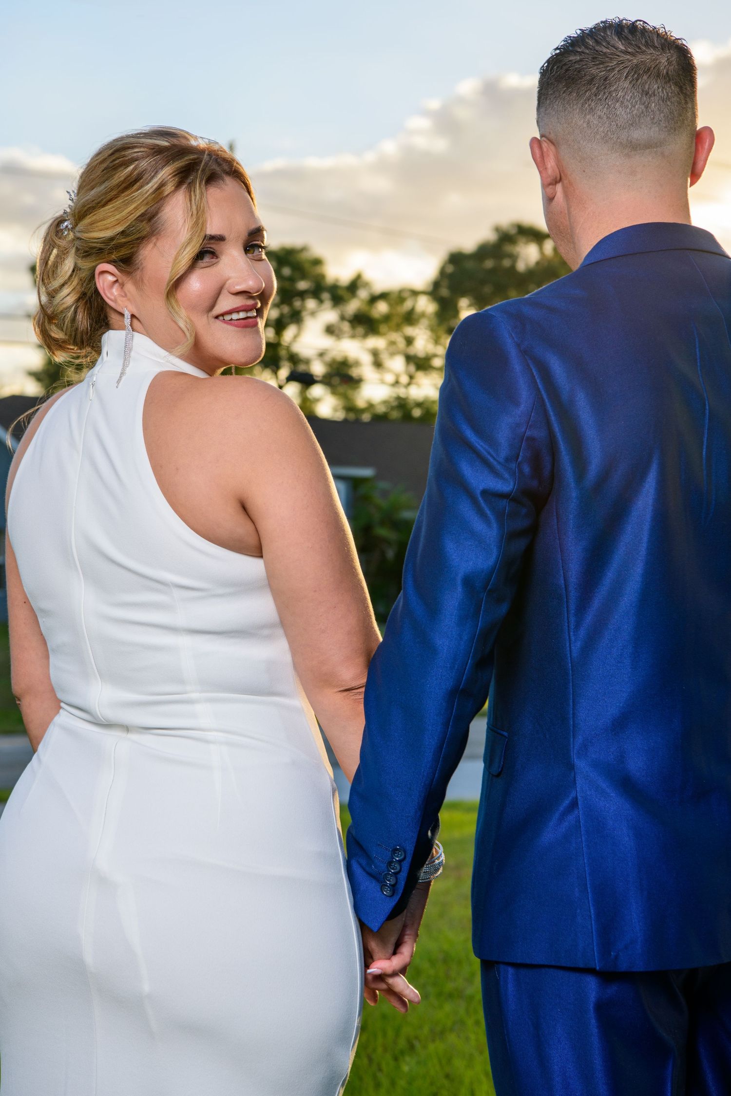 Wedding couple in white dress and blue suit embracing outdoors at sunset.