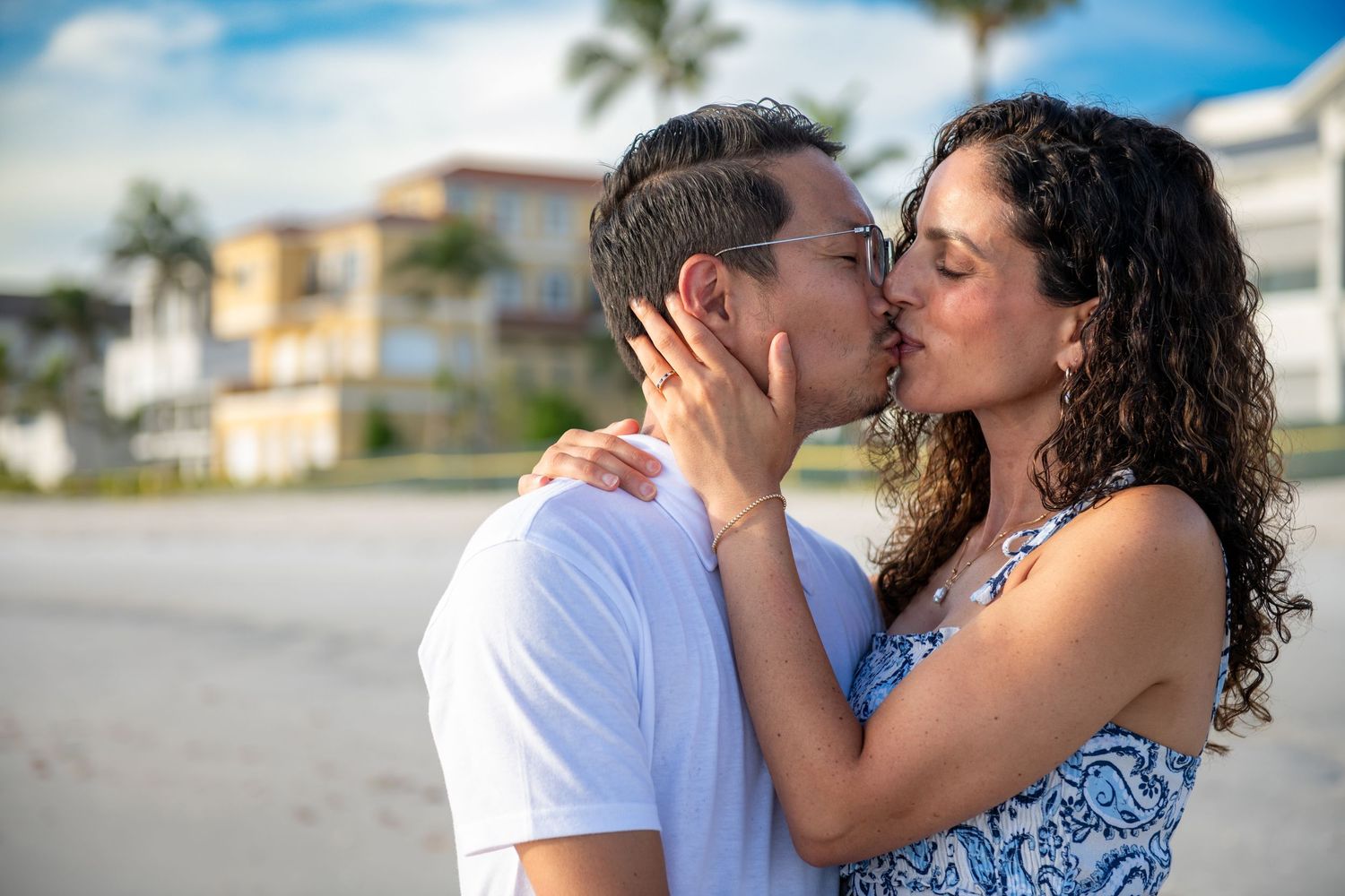 A romantic moment captured between a couple on a sunny beach with palm trees and buildings visible.