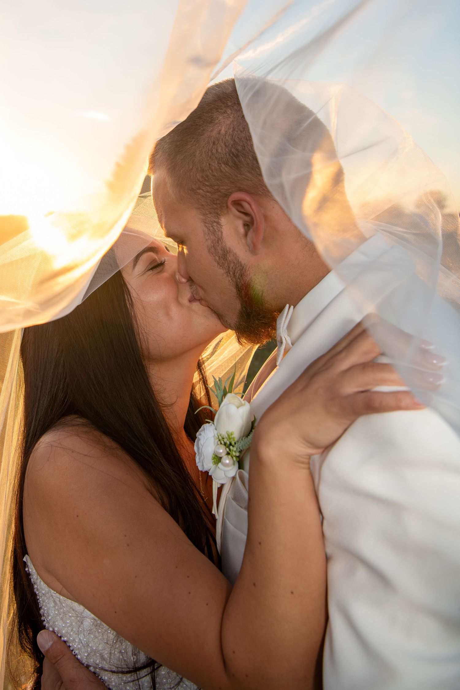 A romantic kiss between newlyweds backlit by golden sunset light with flowing veil. Wedding photography.
