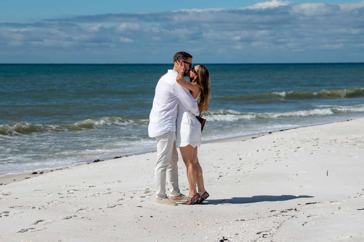 A romantic couple embraces on a white sandy beach with turquoise ocean waters in the background.