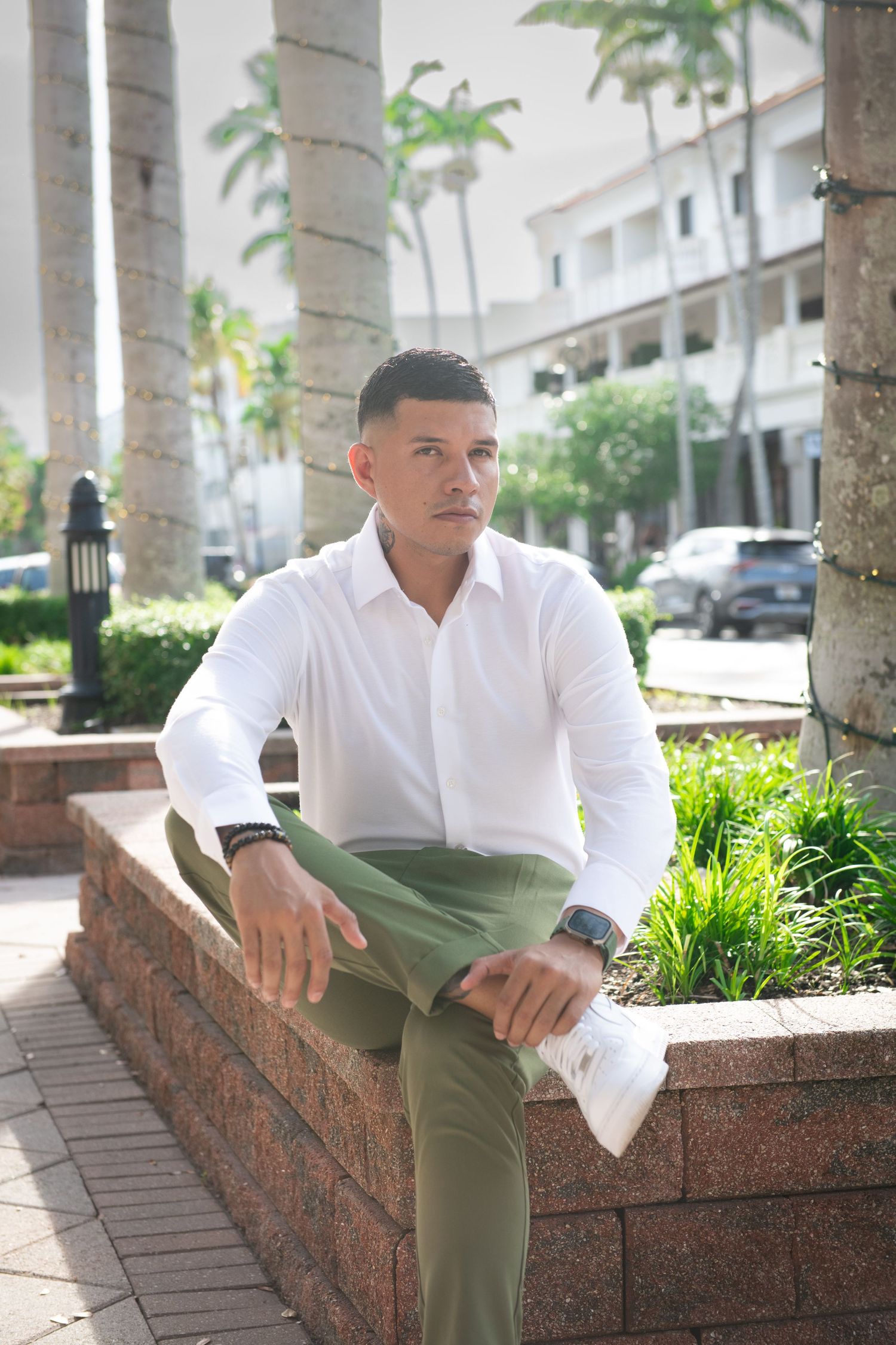 Casual business portrait in white shirt and olive pants seated on brick wall with palm trees in background.