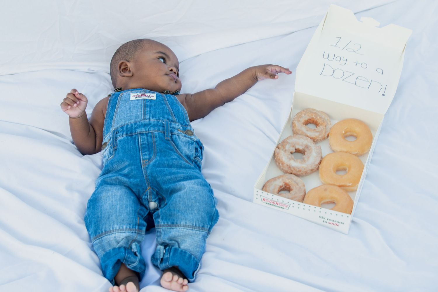 Baby in denim overalls lies next to a book on white bedding.