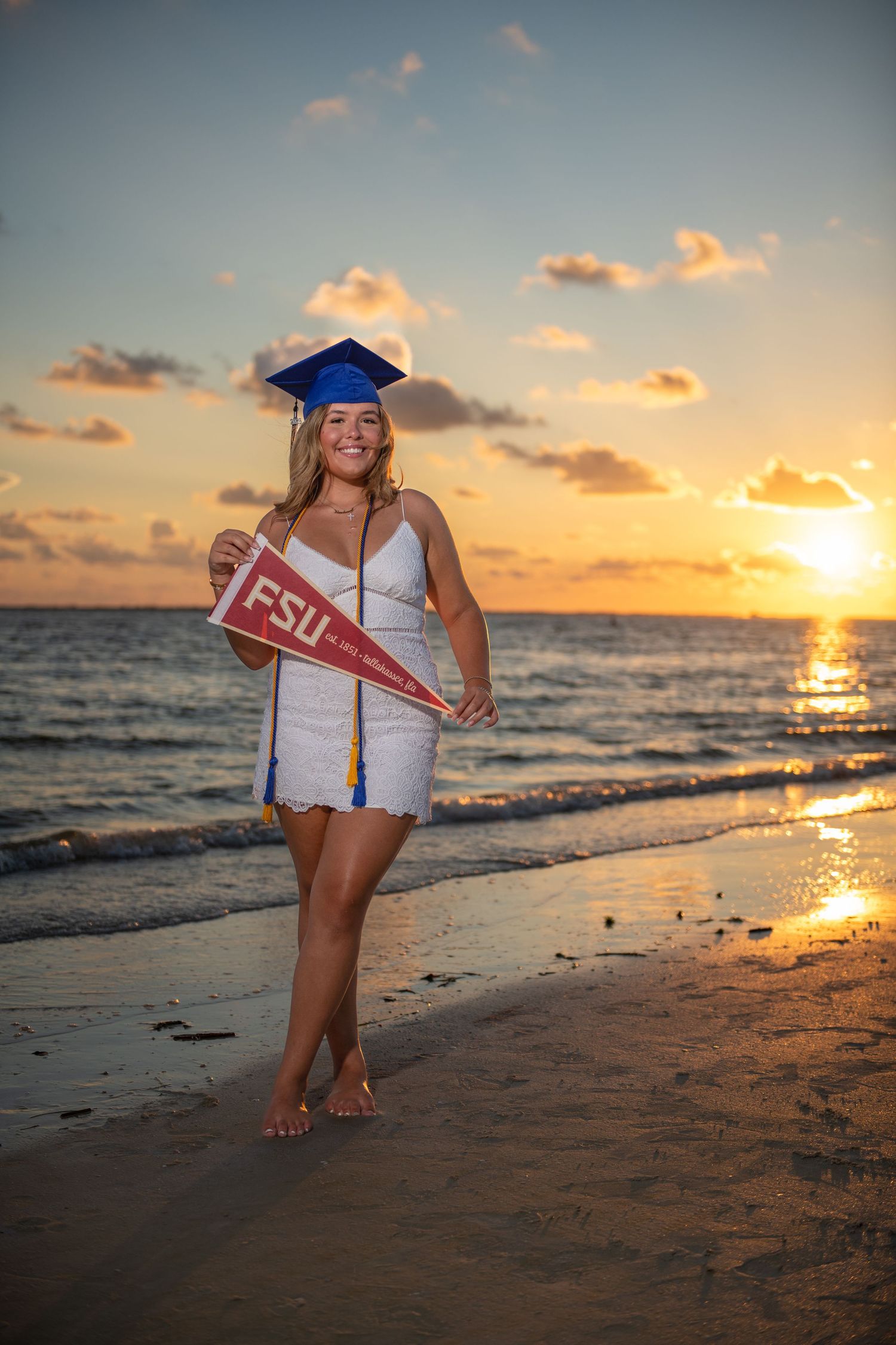 FSU graduate in white dress and blue cap poses with pennant on beach at sunset.
