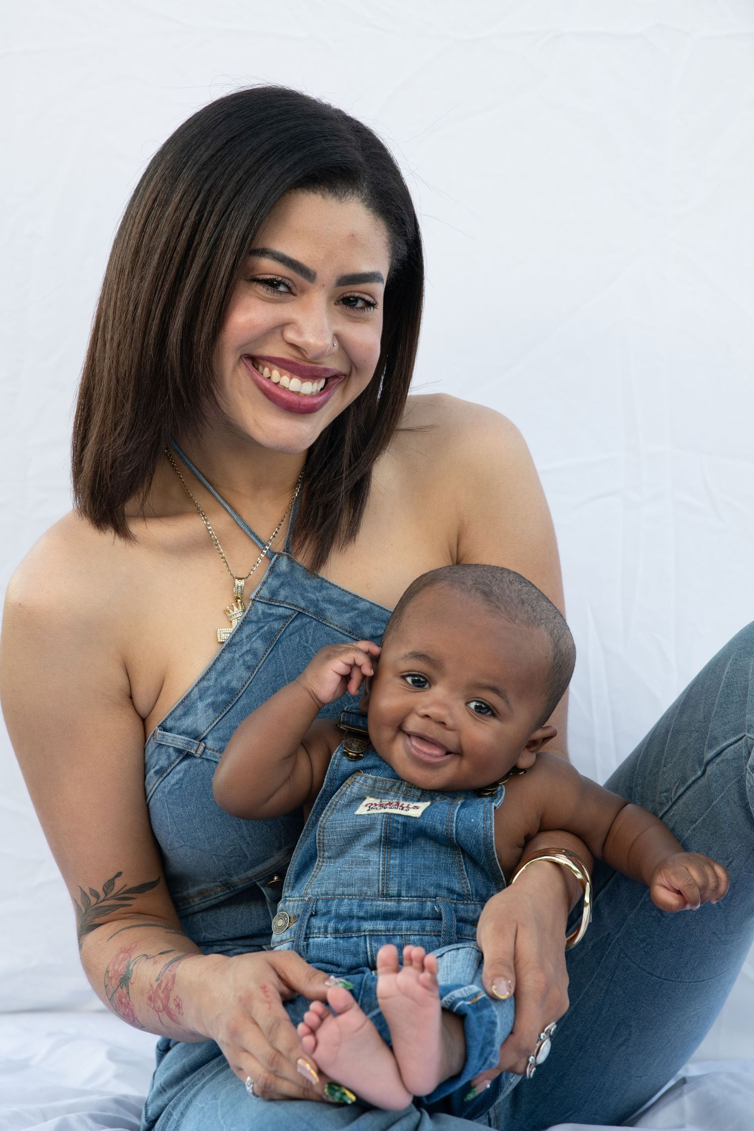 Baby in denim overalls smiles while being held against white backdrop.