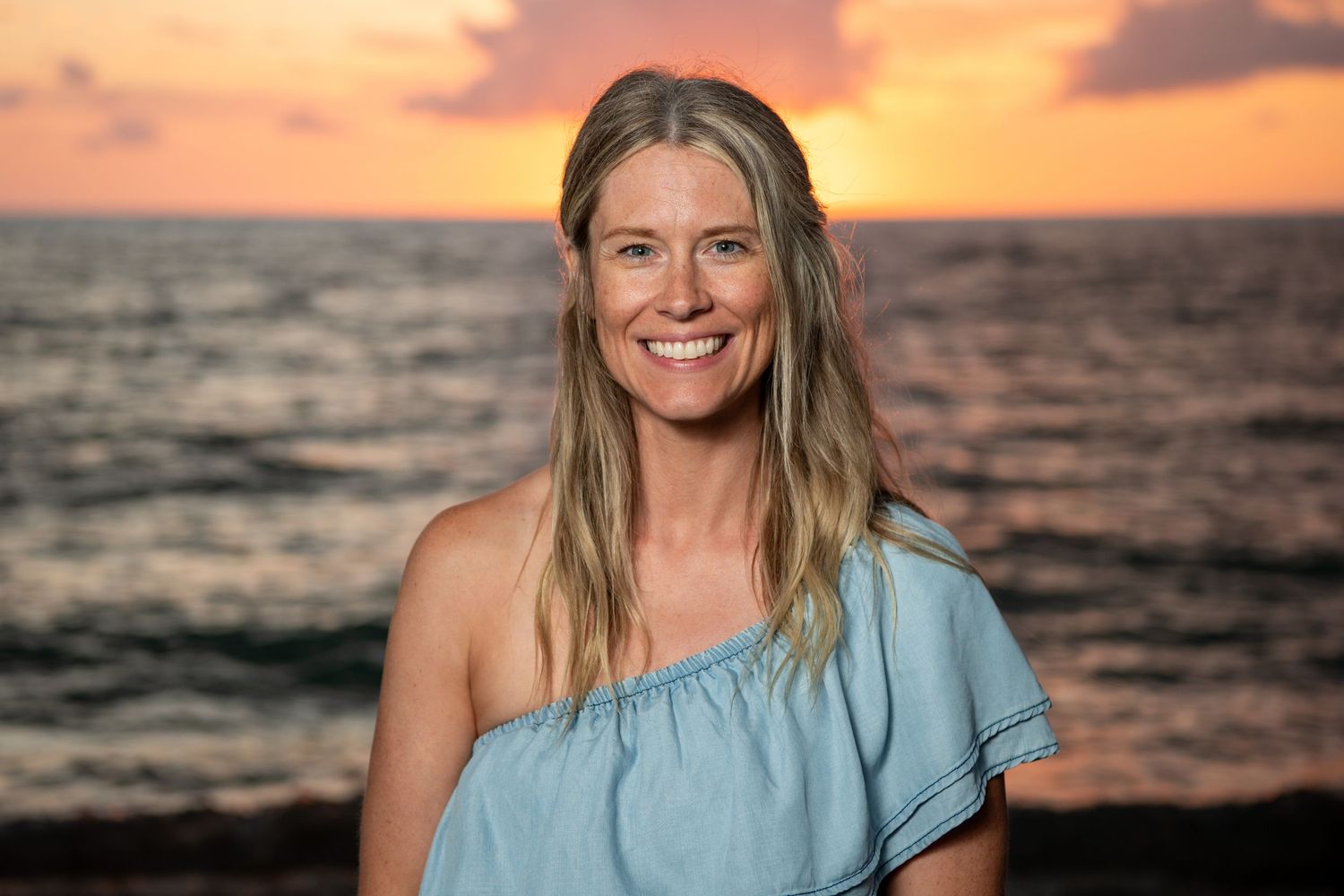 Woman in light blue off-shoulder top poses against sunset ocean backdrop. Beach photoshoot.