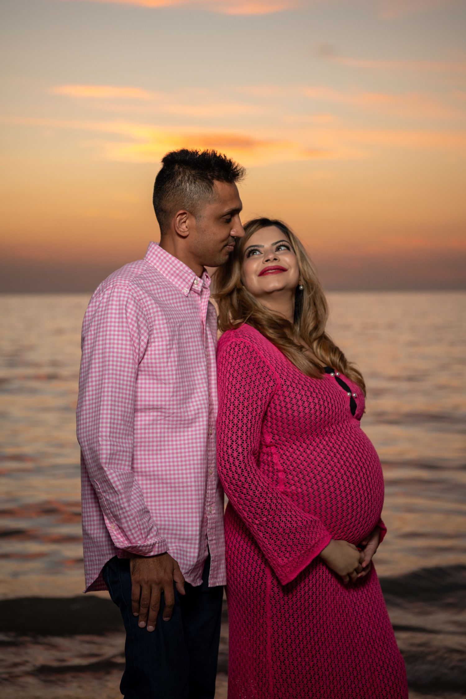 A romantic sunset beach photo of an expectant couple with the mother wearing a pink sweater dress. Maternity photoshoot. Couples photos.