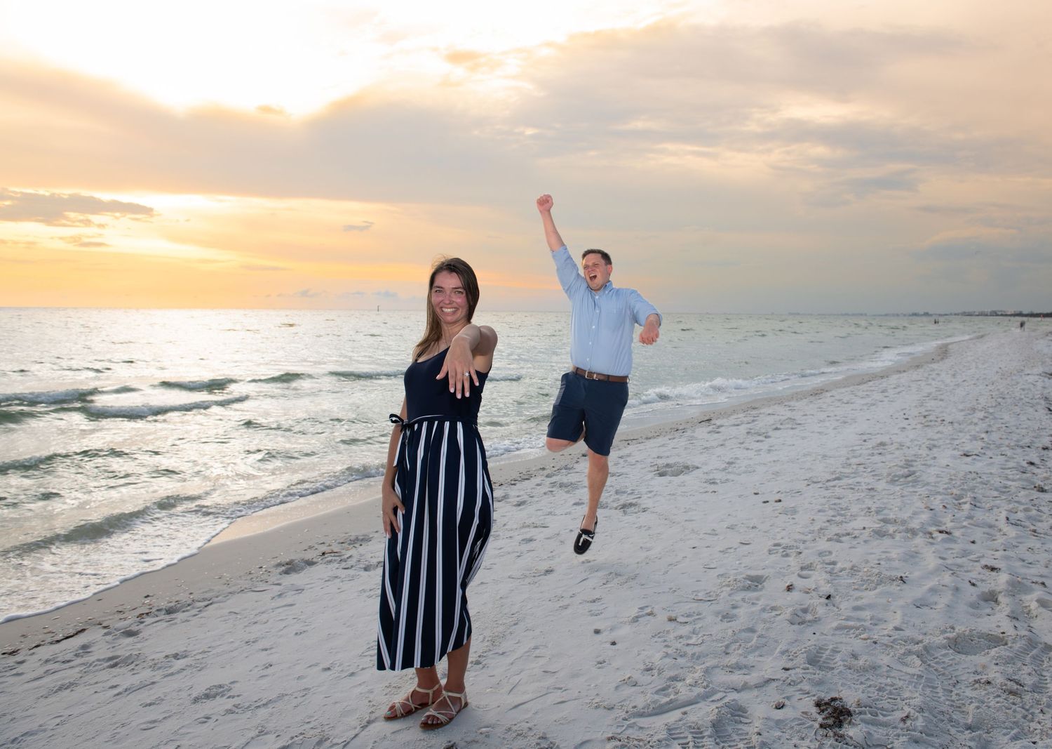 A couple walks along a white sandy beach at sunset with dramatic orange clouds in the sky.