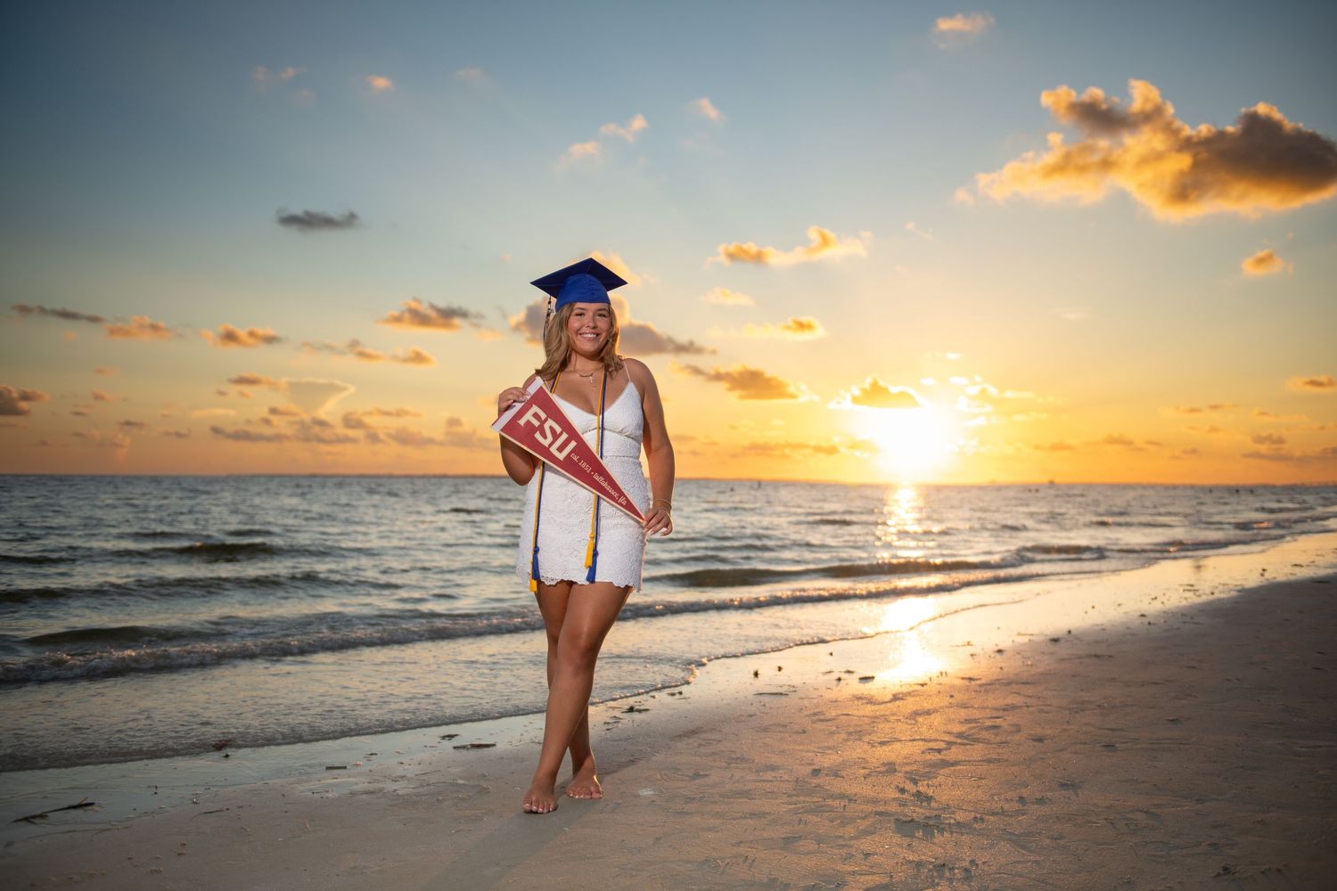 Graduate in cap and gown poses on beach at sunset holding FSU pennant.