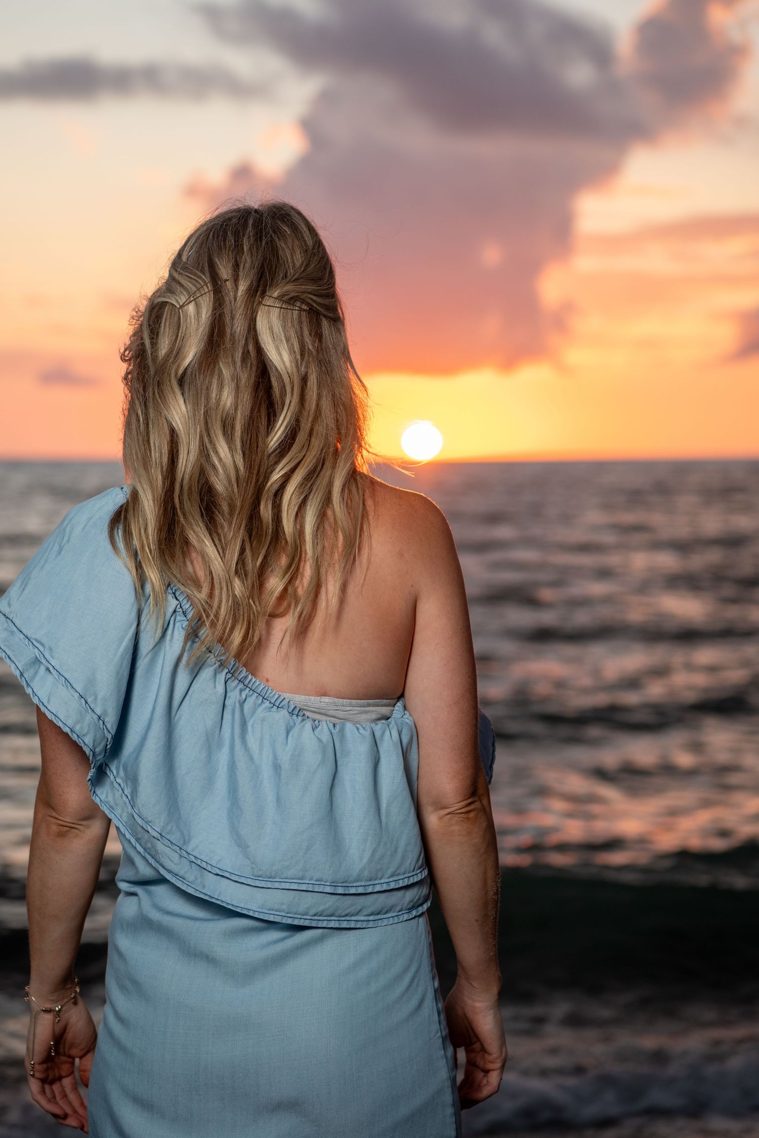 A woman in a blue ruffled top poses against a dramatic sunset ocean backdrop. Beach photoshoot. Family photoshoot,