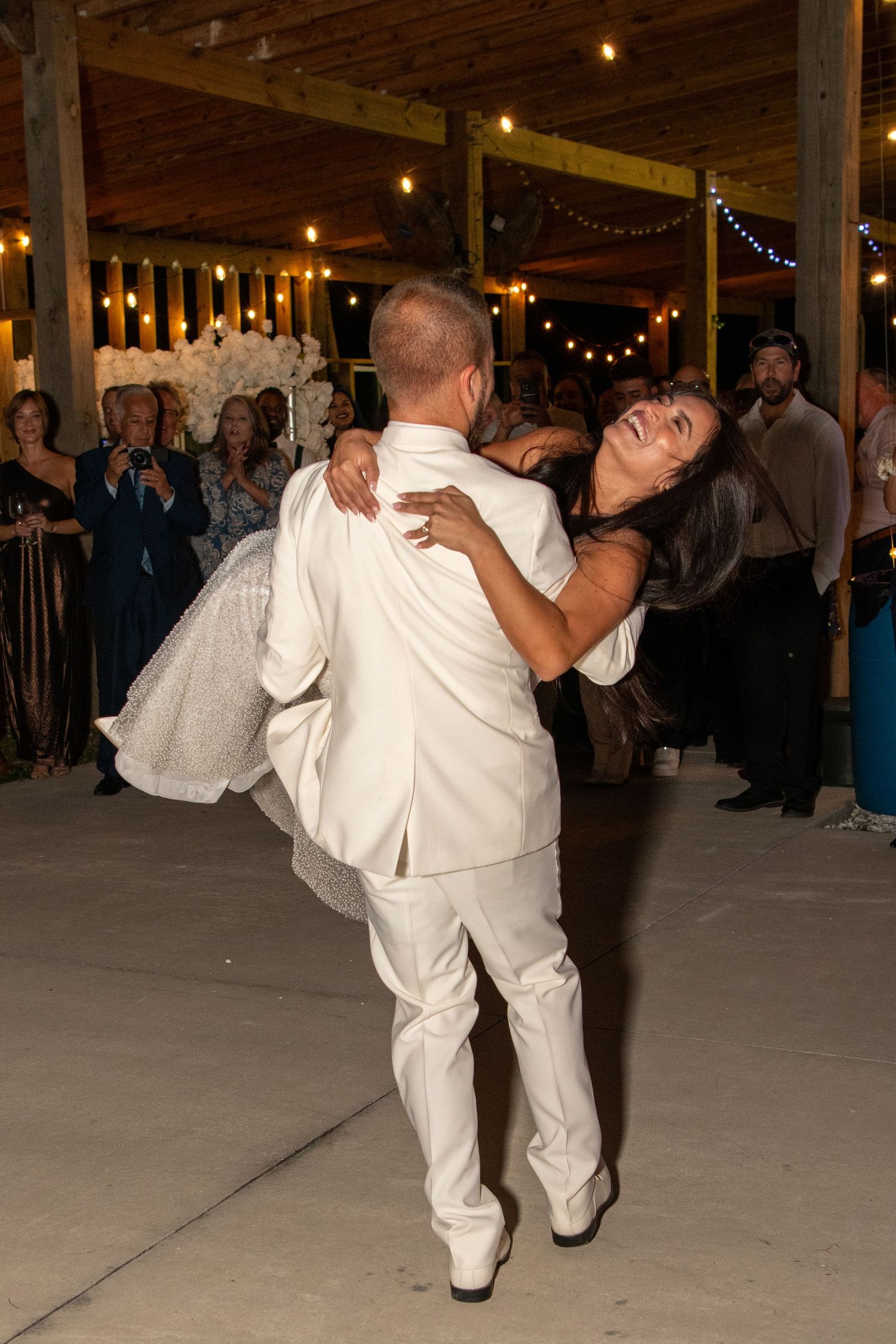 Couple performs romantic dance dip under string lights at indoor reception venue. Wedding photography.