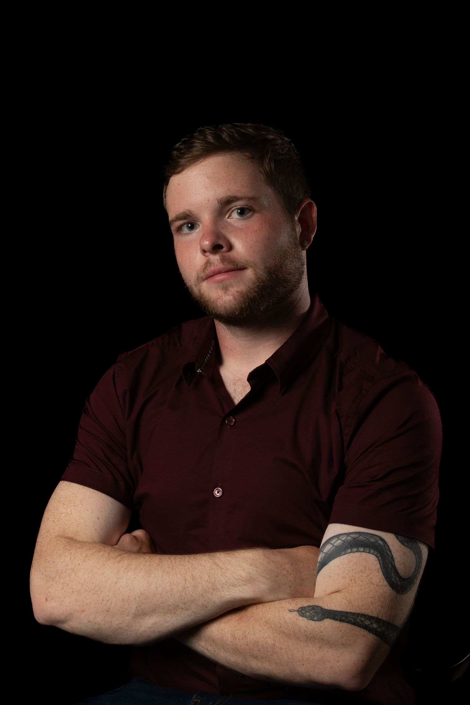 Studio portrait against black background featuring burgundy polo shirt and visible tattoos.