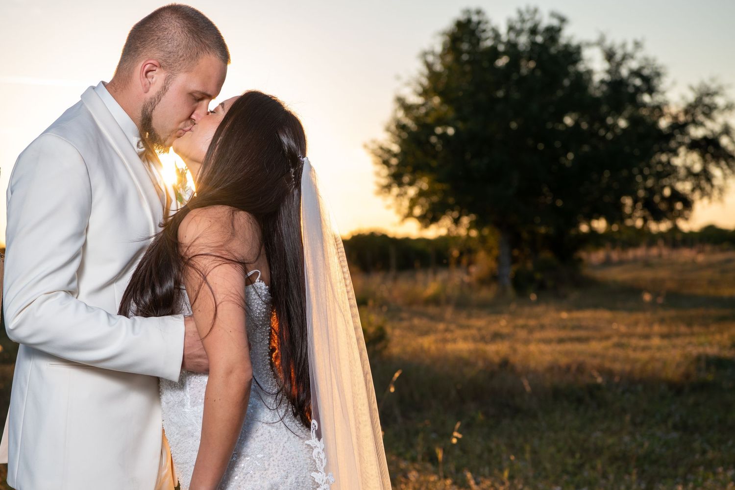 Newlyweds share an intimate moment at sunset in a field with trees in the background. Wedding photography.