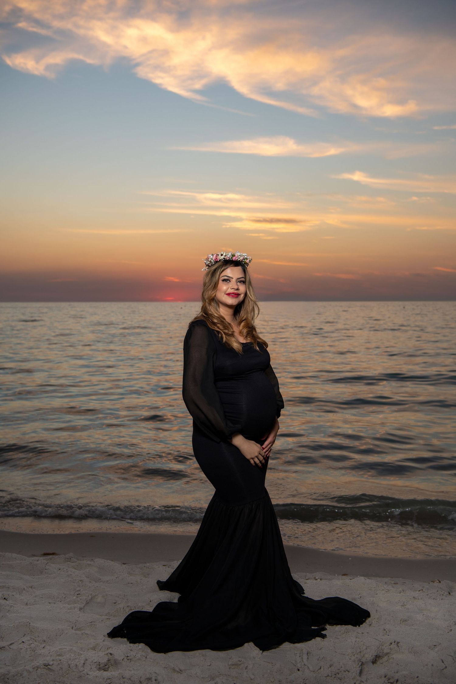 A maternity photoshoot featuring a woman in a flowing black dress posing on the beach at sunset. Maternity photoshoot. Couples photos.