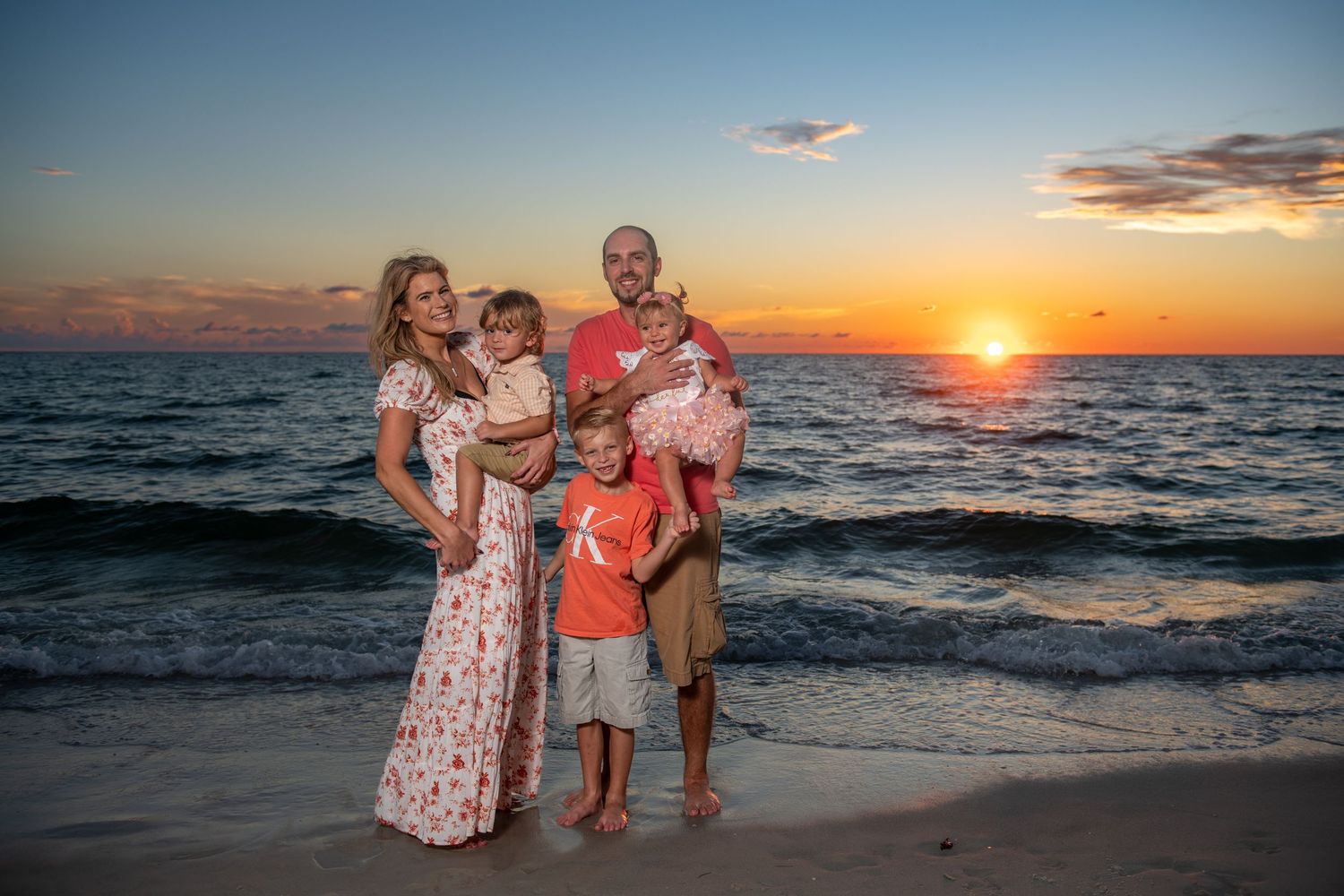 A family poses together on the beach during a dramatic orange sunset over the ocean.