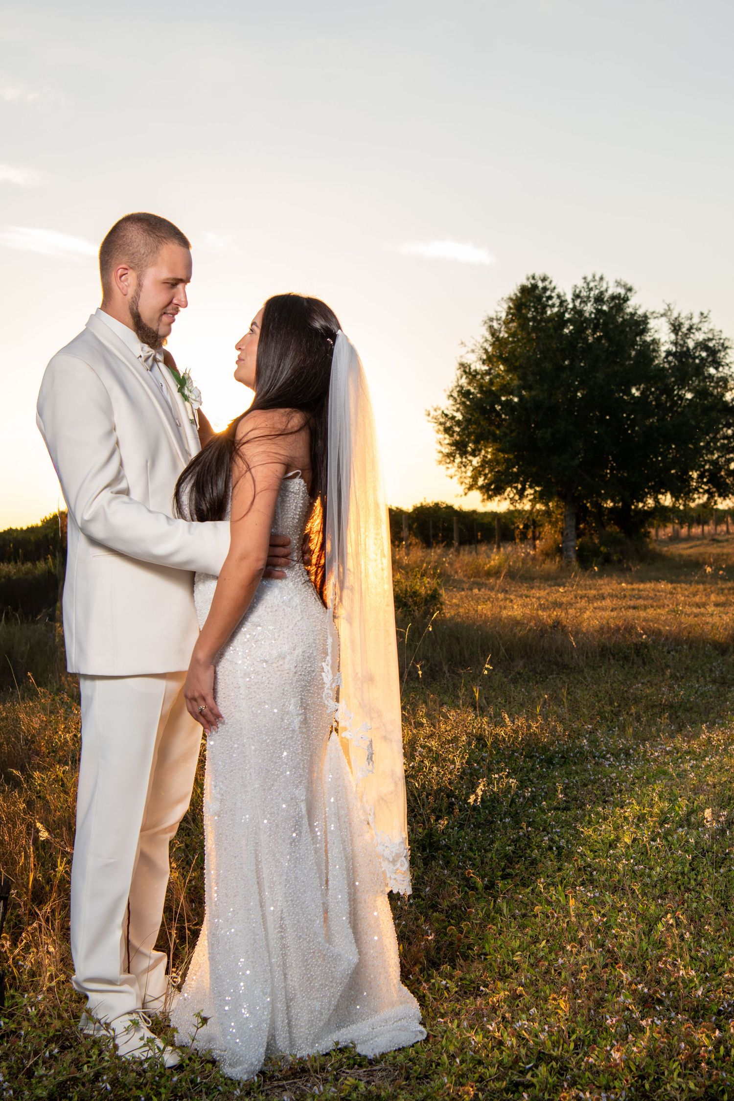 A couple in wedding attire poses together in a field during golden hour sunset. Wedding photography.