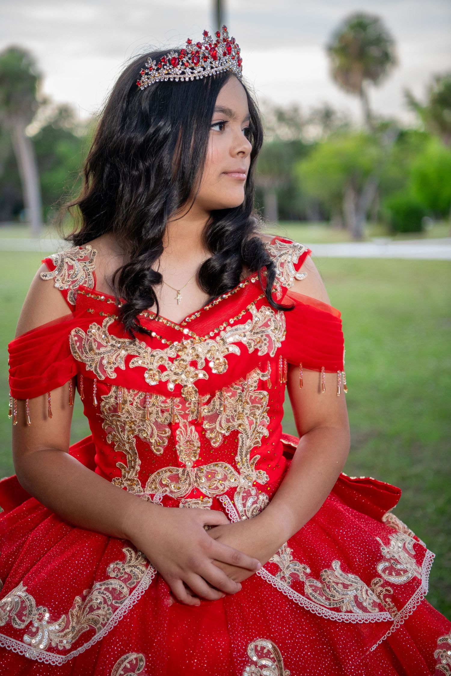 Elegant red and gold embroidered formal dress photographed outdoors in a garden setting. Quinceañera photoshoot.