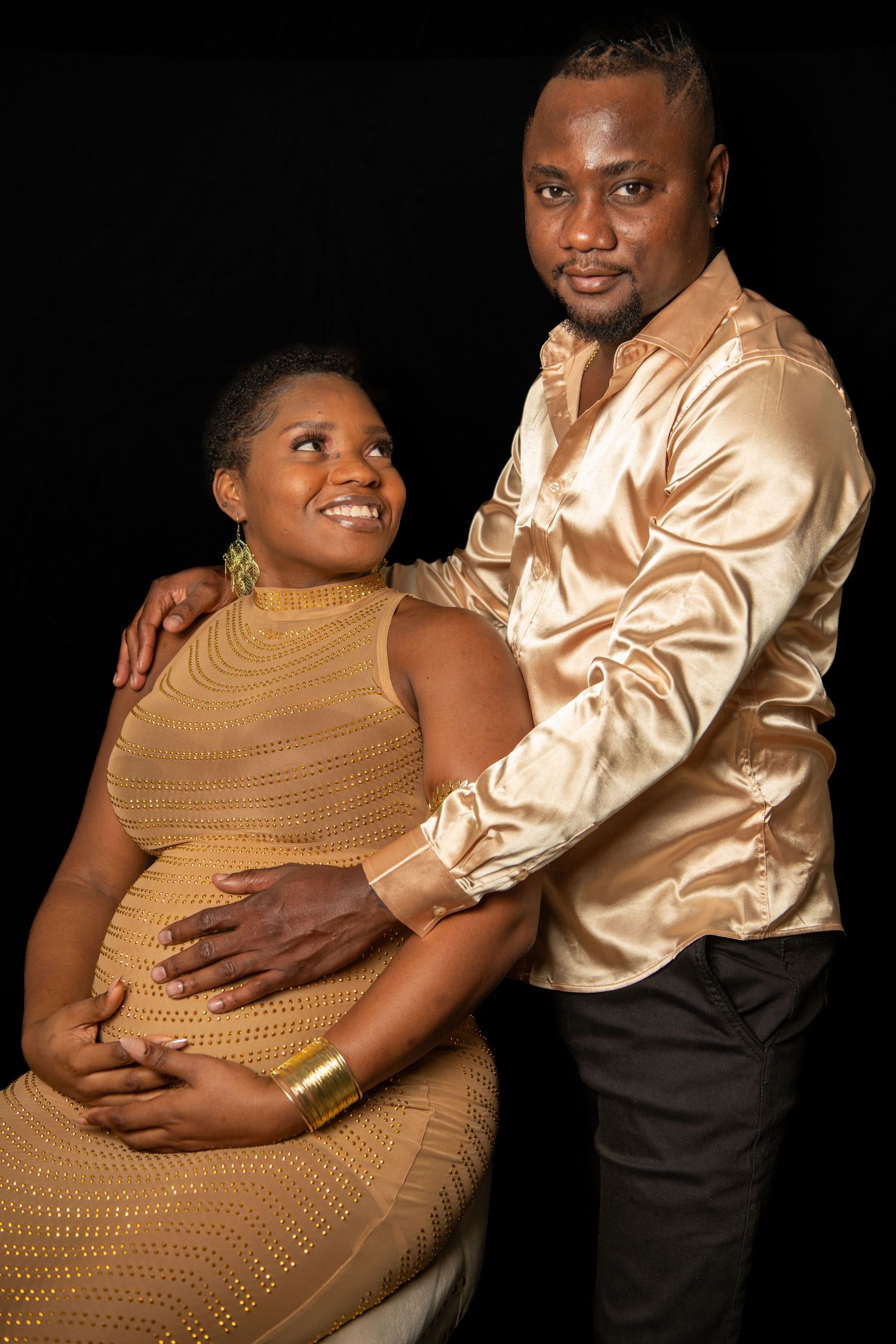 Studio maternity portrait with couple in gold formal attire against black background. Maternity photoshoot.