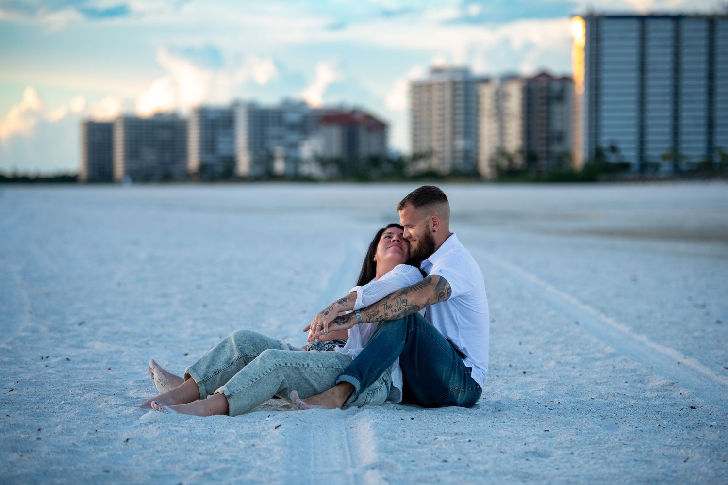 Couple sitting together on sandy beach at sunset with city skyline in background. Maternity photoshoot.