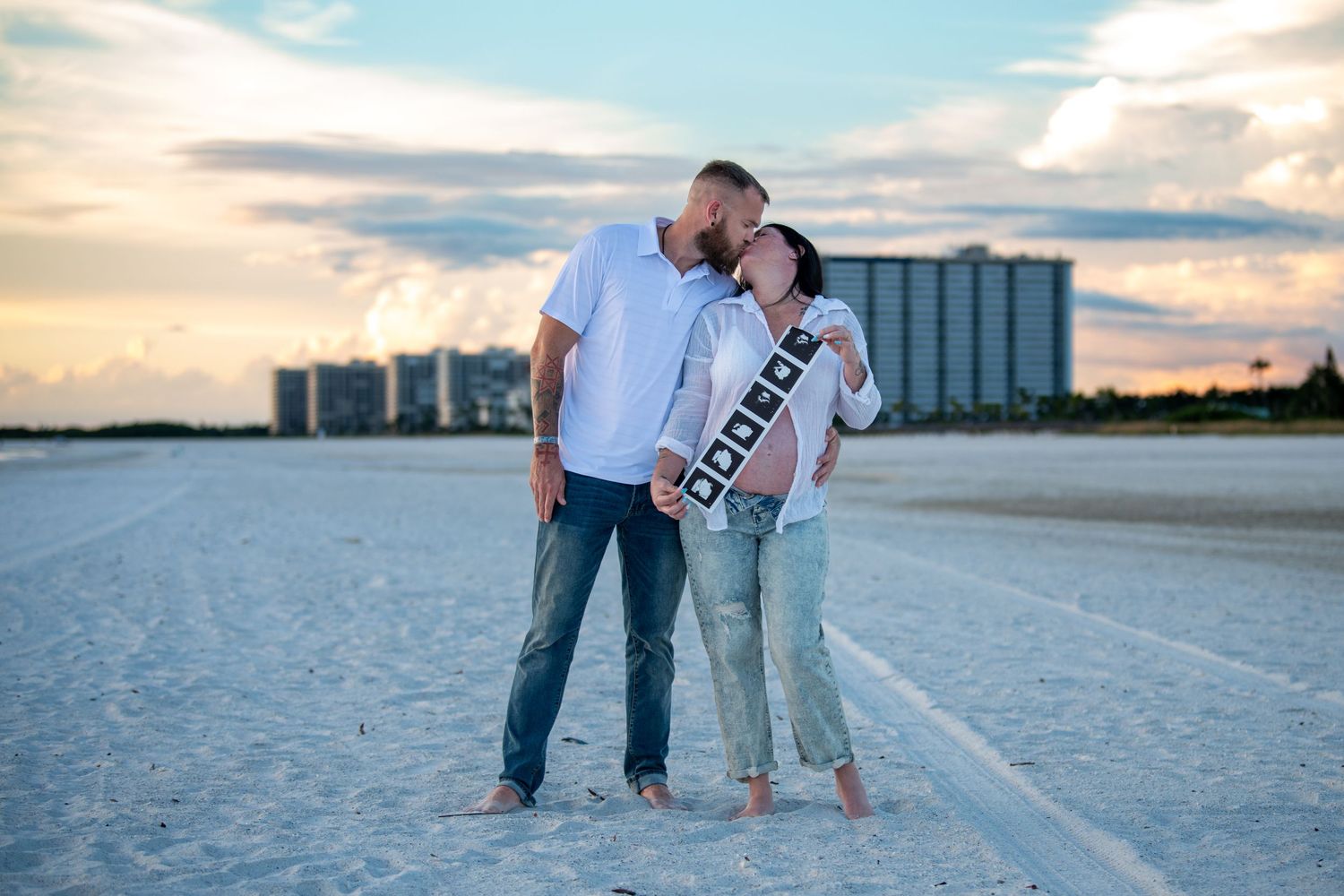 Couple embracing on beach at sunset with coastal buildings in background. Maternity photoshoot.