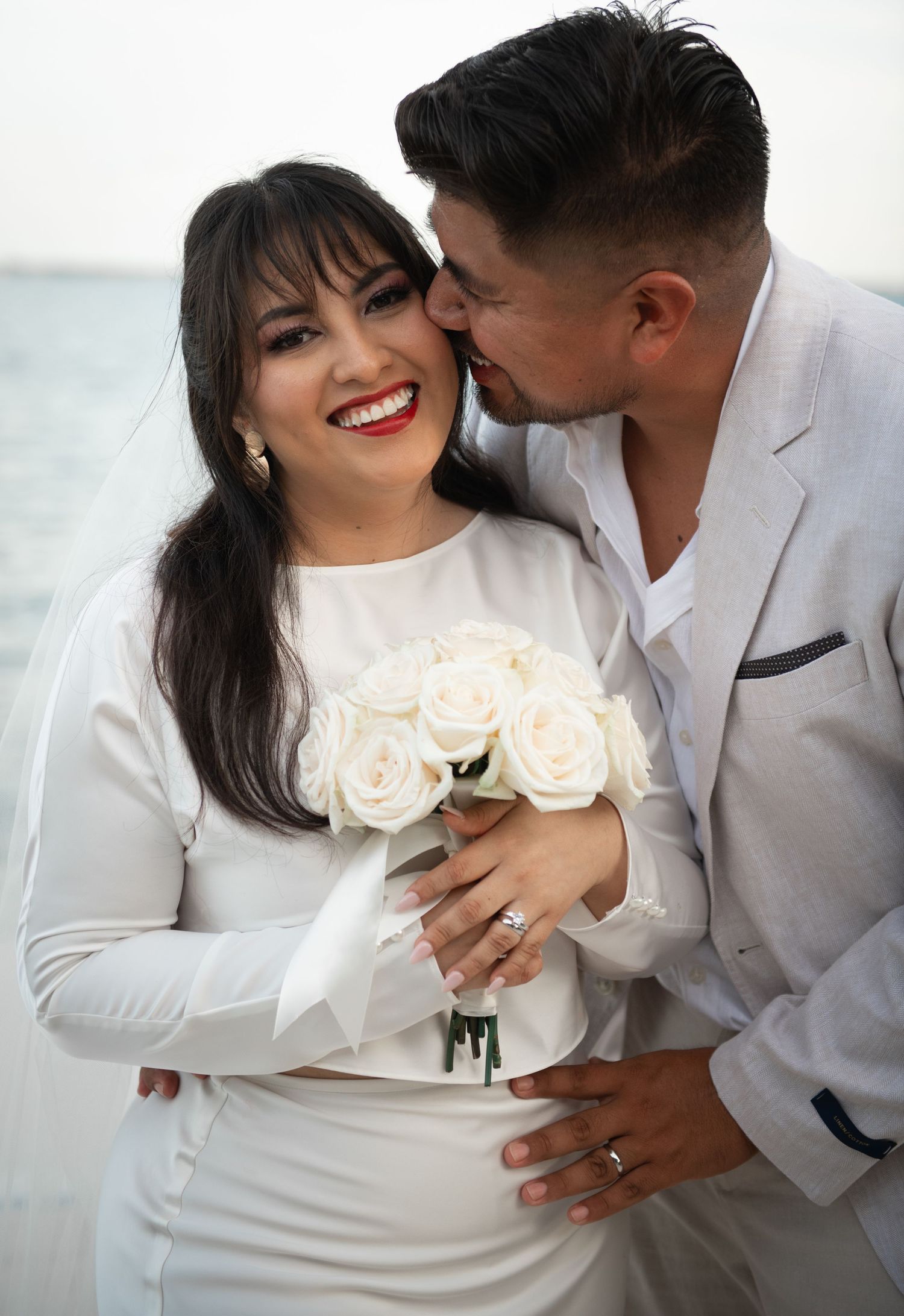 Romantic couple in white attire sharing intimate moments with cream roses against bright background.