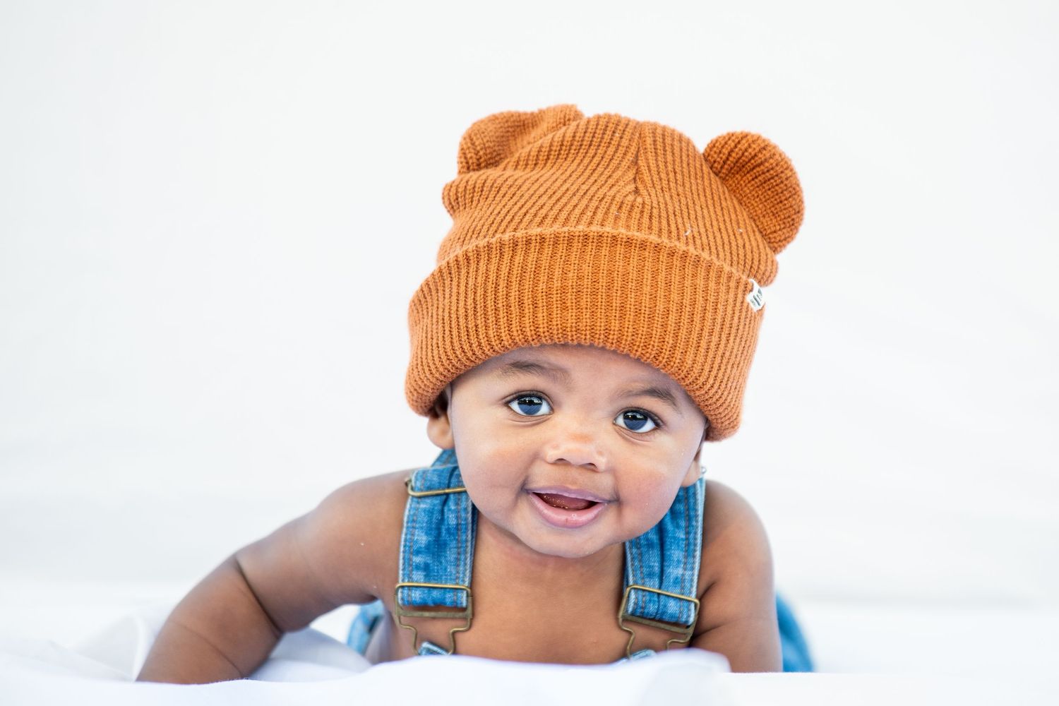 Baby wearing orange knit beanie and denim overalls smiles at camera.