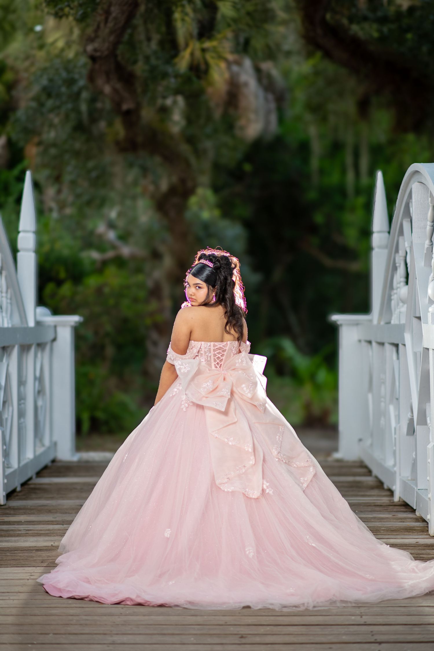 A flowing pink quinceañera dress photographed on a white wooden bridge in a garden setting. Quinceañera photos.