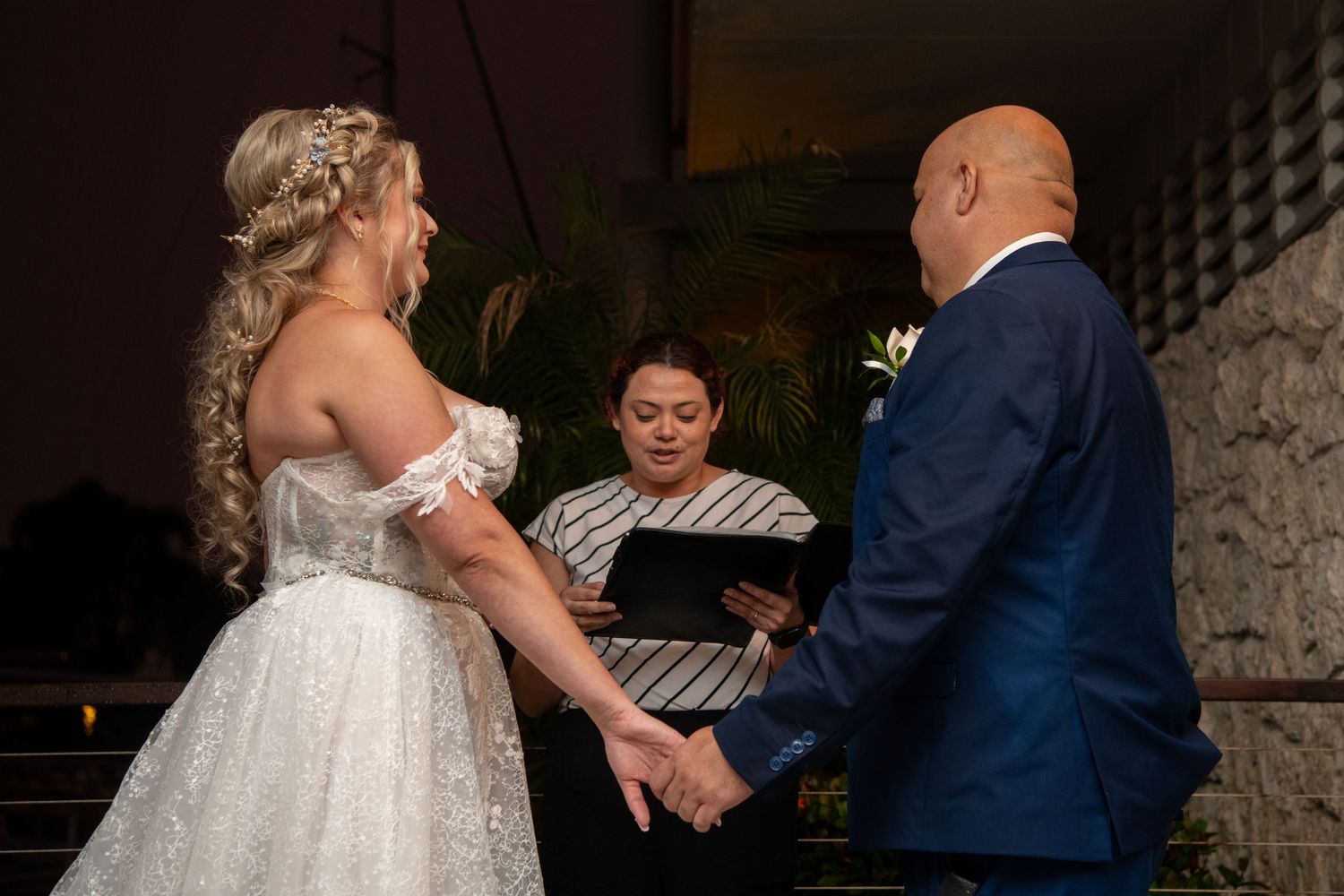 A couple exchanges rings during an indoor wedding ceremony with palm trees visible in background.