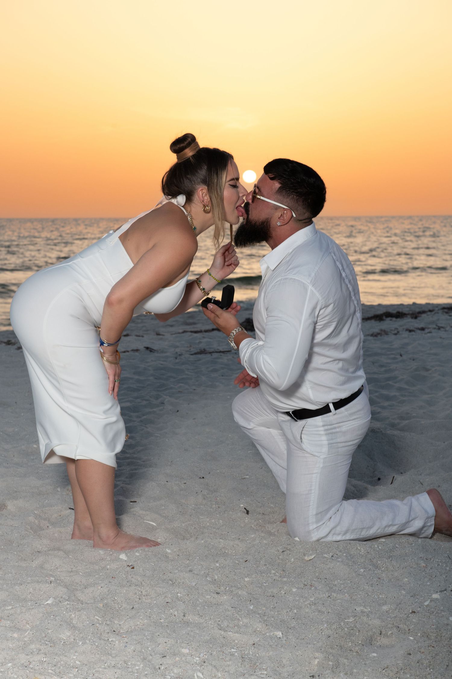 Beach proposal scene at sunset with person kneeling in white attire on sandy shore.