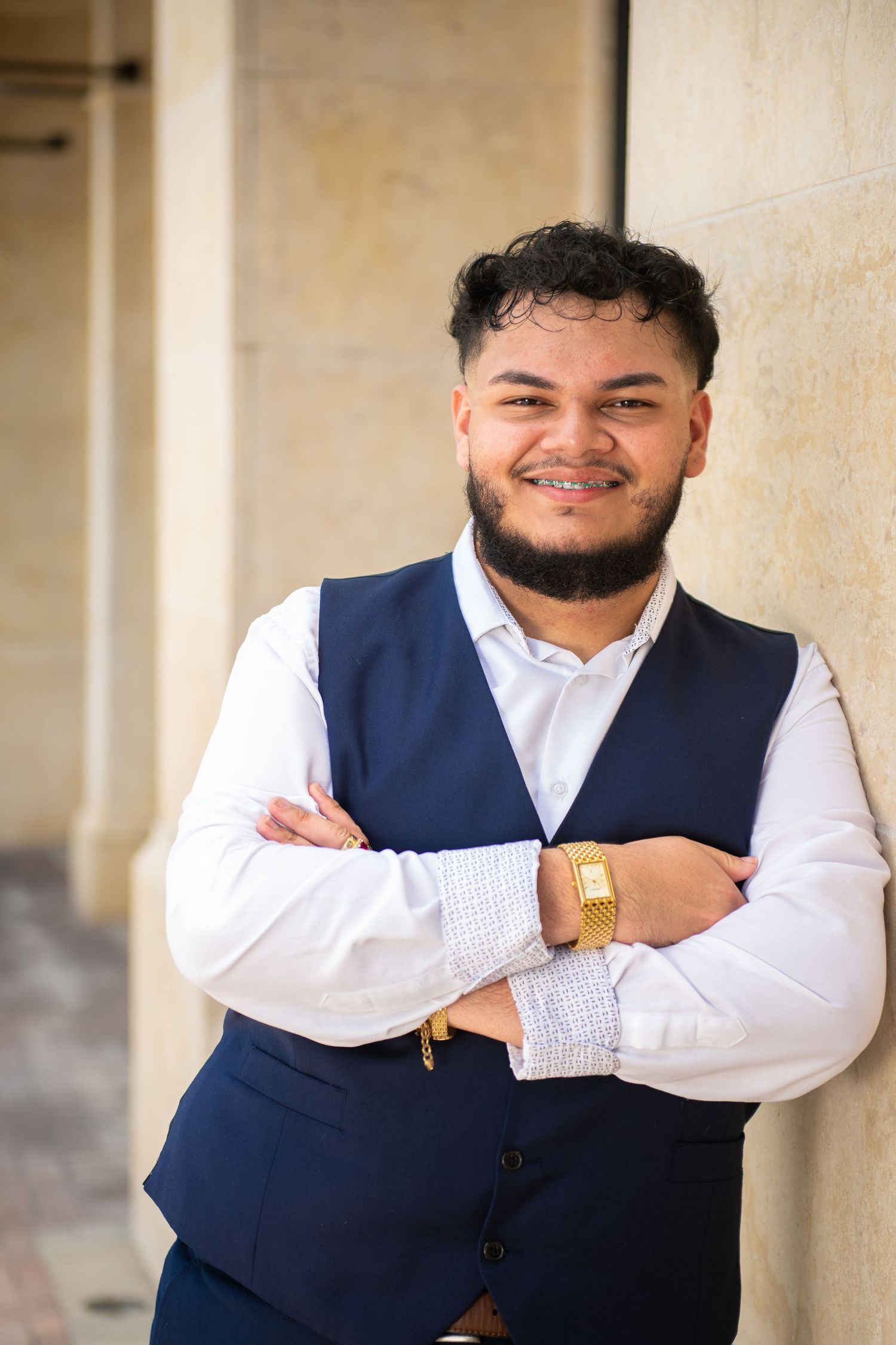 Formal portrait in navy vest and white shirt showing confident pose against beige wall.