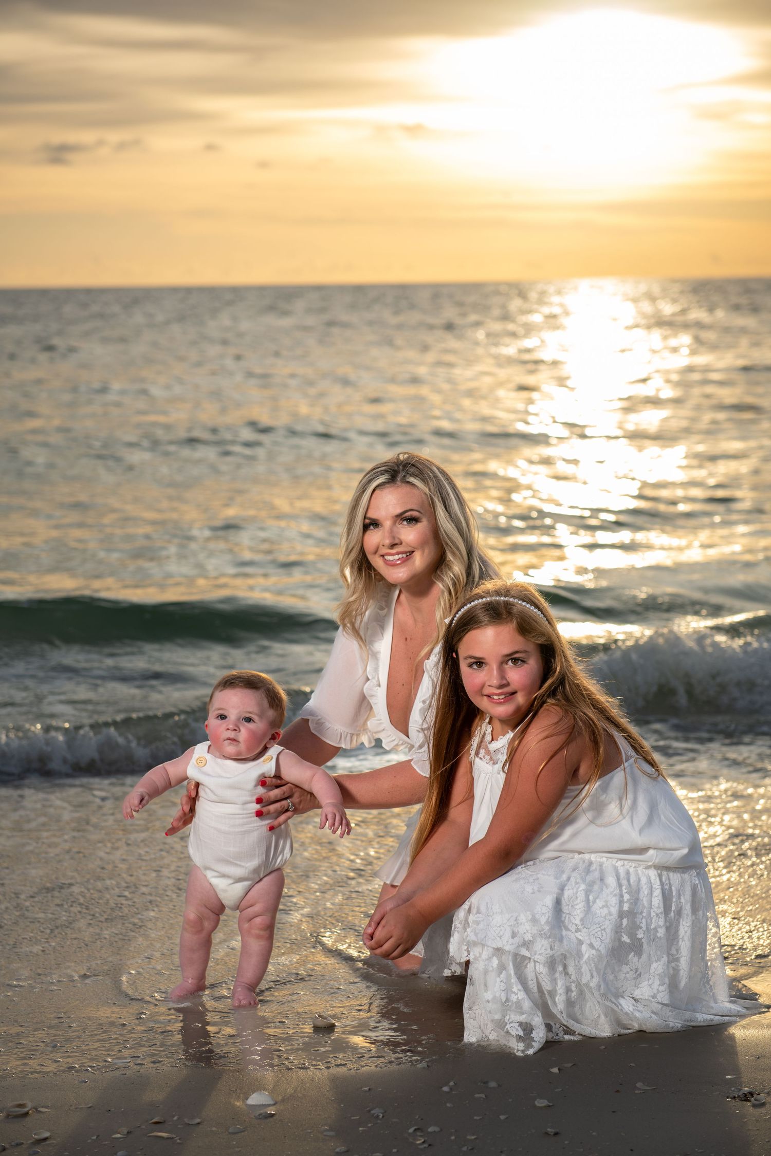 Three people in white dresses play with baby at beach during sunset.