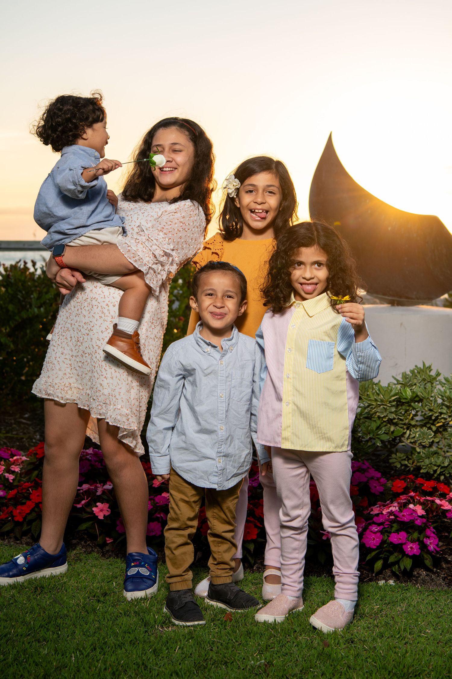 A family group poses together at sunset in front of pink flowers and a curved outdoor sculpture. Family photoshoot.
