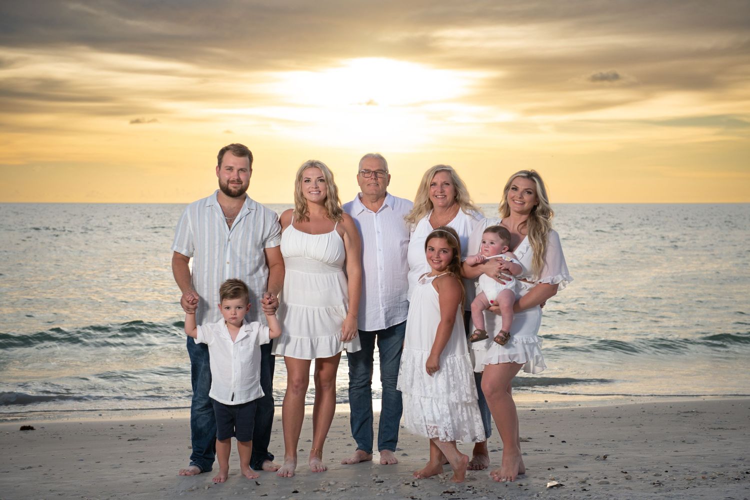 Extended family wearing white outfits poses for beach portraits at sunset.