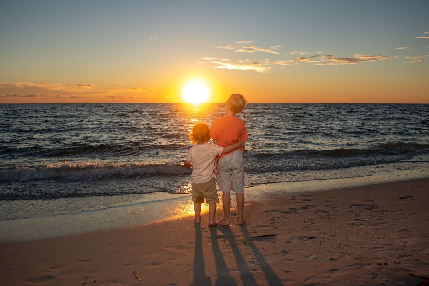 Two silhouettes stand watching a vibrant orange sunset over the ocean.