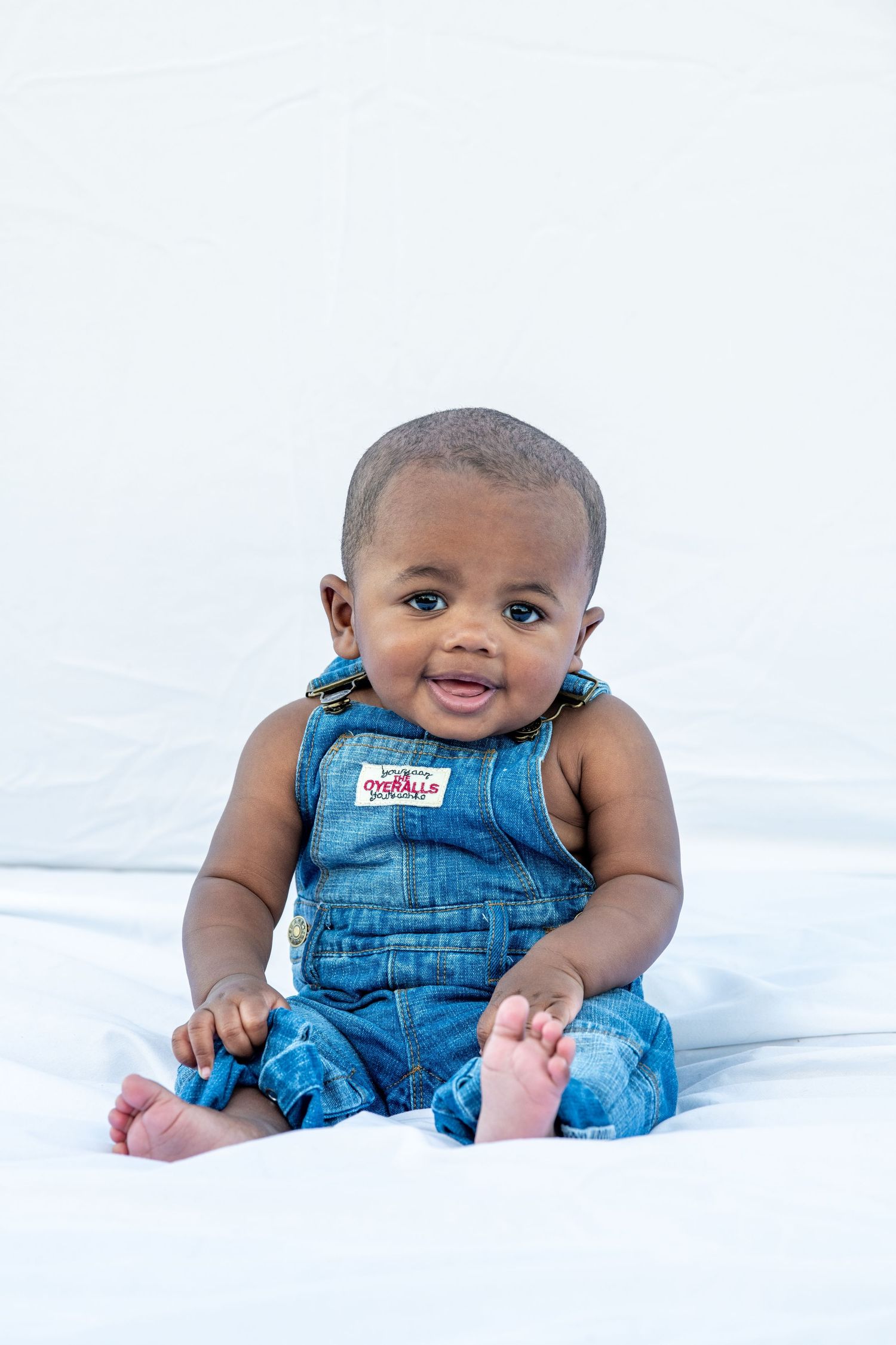 Smiling infant in blue denim overalls sits on white background.